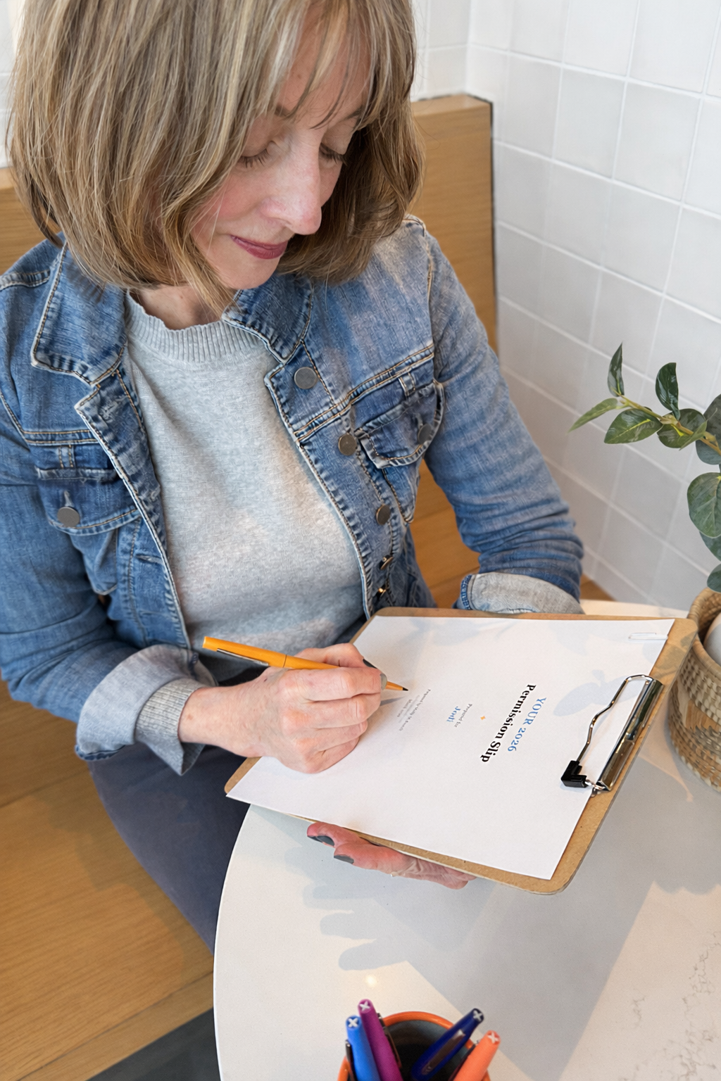 A woman in a denim jacket and gray sweater is sitting at a table, filling out a petition sheet on a clipboard with an orange pen.