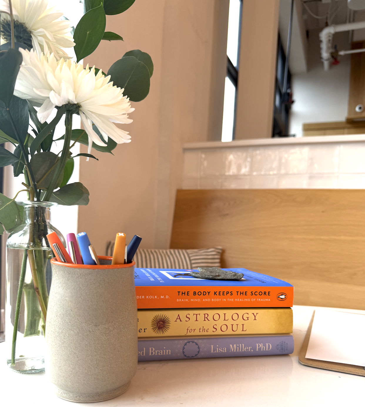A table with a glass vase of white flowers, a beige cup of colorful pens, three stacked books about astrology, brain healing, and trauma, and a closed notebook. In the background, a wooden bench with a striped pillow and a part of a room with windows are visible.