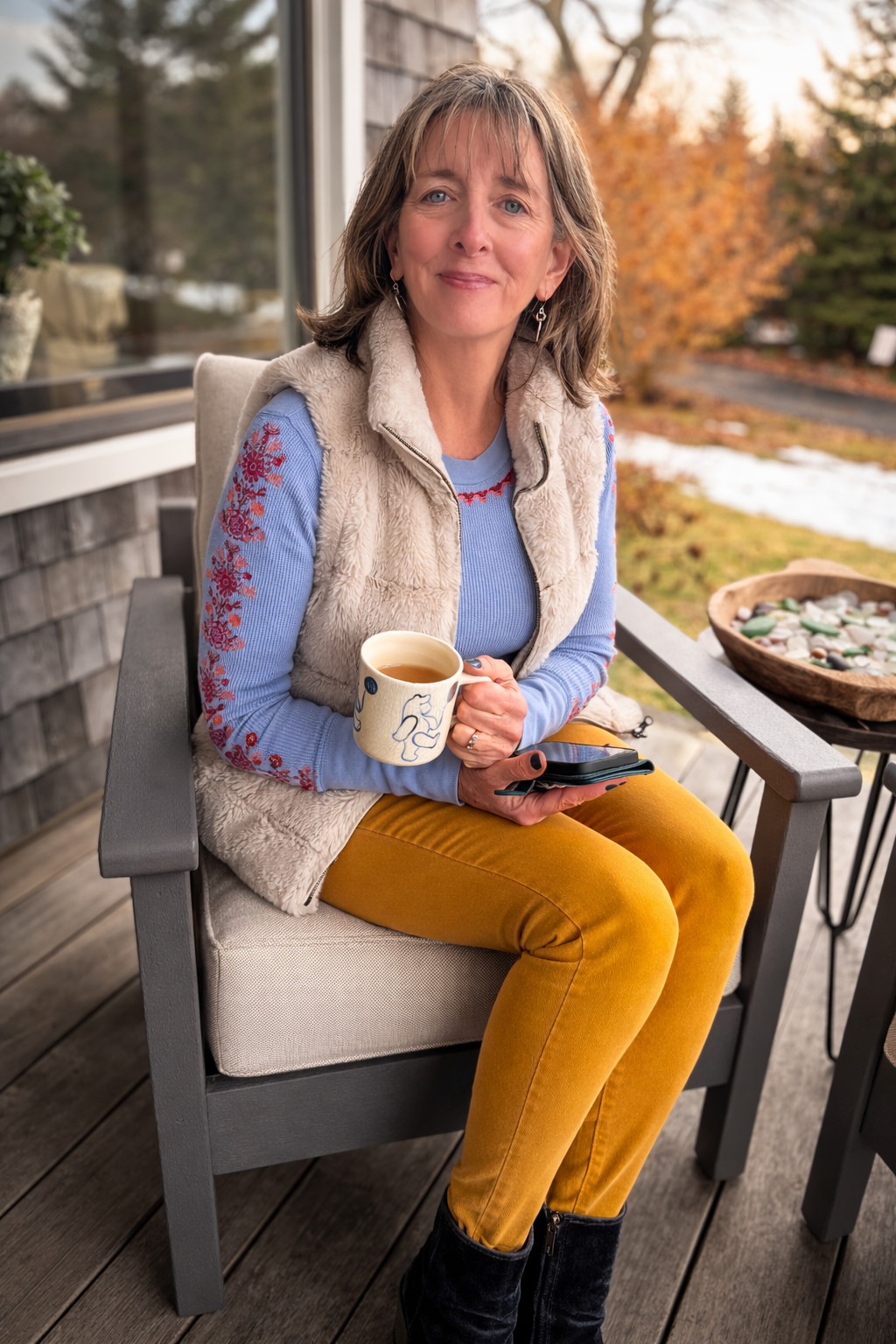 A woman with shoulder-length brown hair and blue eyes sitting on a patio chair outdoors, holding a cup of tea or coffee, wearing a blue sweater, a beige vest, mustard yellow pants, and black boots, with a tray of rocks or stones on a side table nearby. In the background, there are autumn trees with orange leaves and a partly snow-covered ground.
