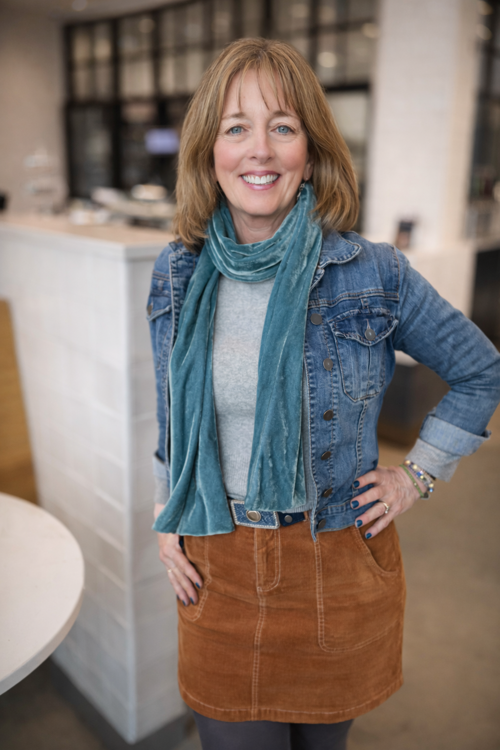 Woman with shoulder-length reddish hair, wearing a denim jacket, teal scarf, gray top, burnt orange skirt, and bracelets, standing indoors with a blurred background.