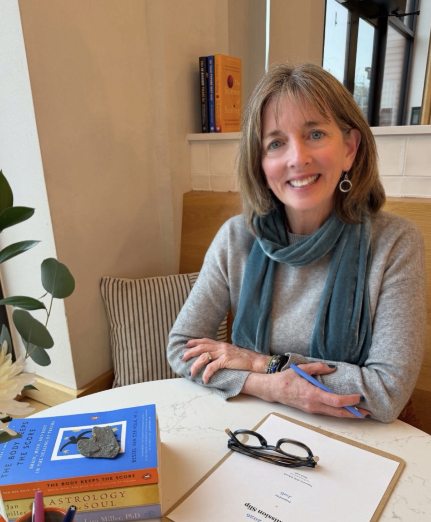 A woman with shoulder-length brown hair and blue eyes smiling, seated at a table with books, glasses, and a pen, in a cozy indoor setting with a window and wall decor in the background.