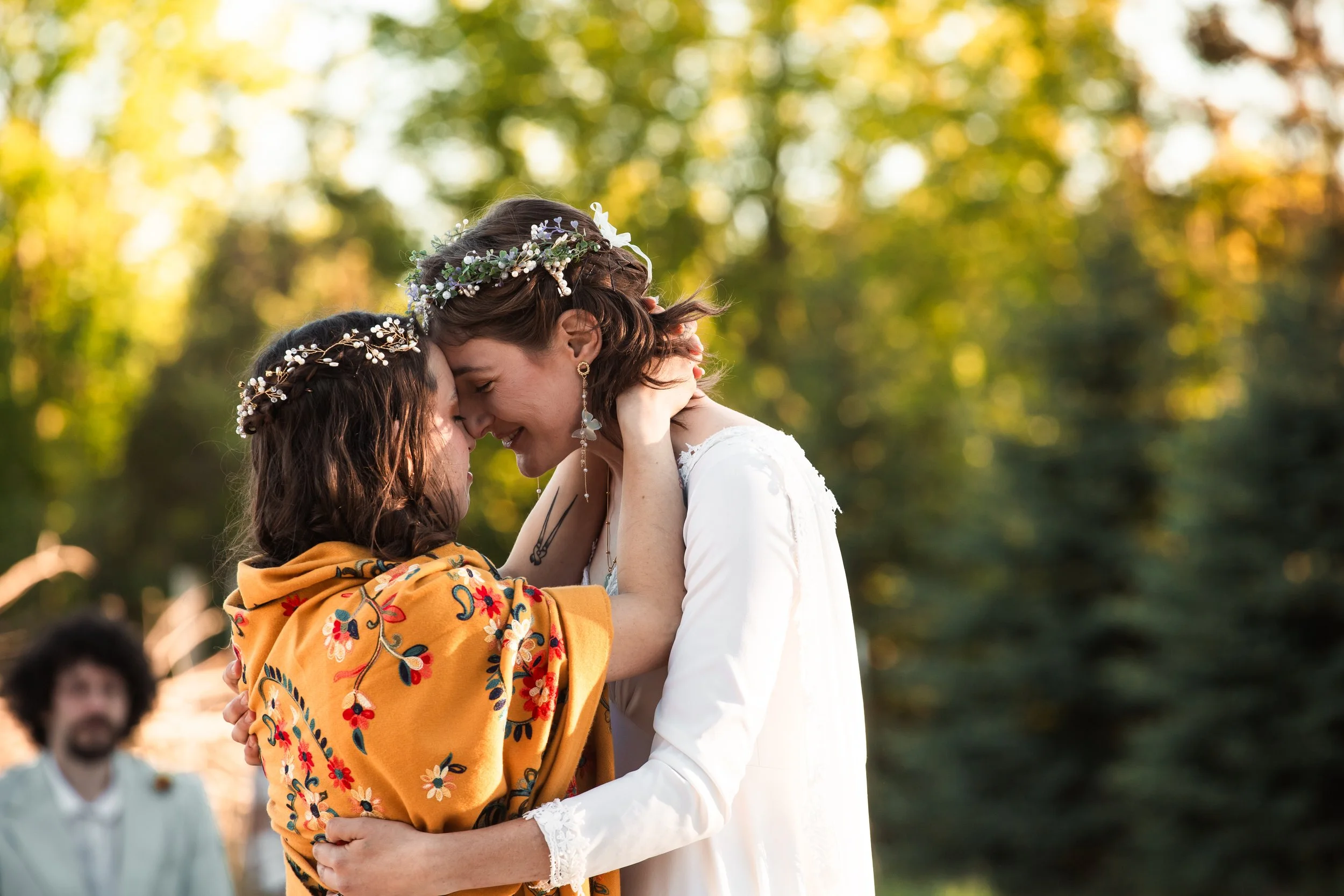 Two women, one in a white dress and the other in a colorful shawl, are embracing each other and touching foreheads in a romantic outdoor setting during what appears to be a wedding or special celebration. They are wearing floral headpieces, with blurred greenery in the background.
