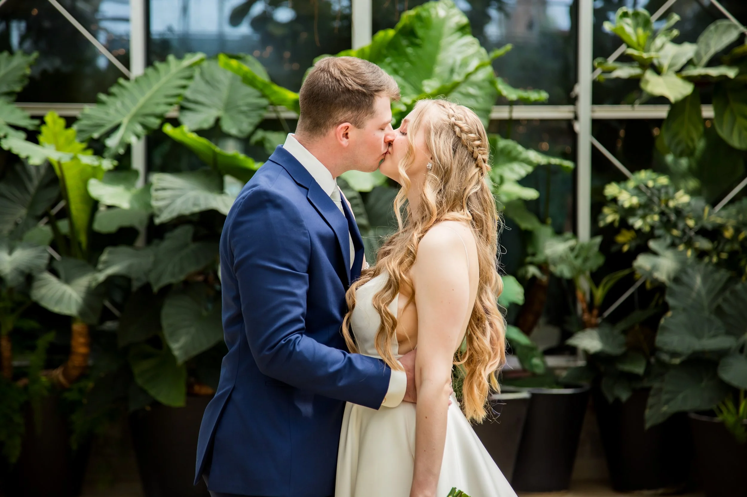A couple in wedding attire sharing a kiss inside a greenhouse with lush green plants.