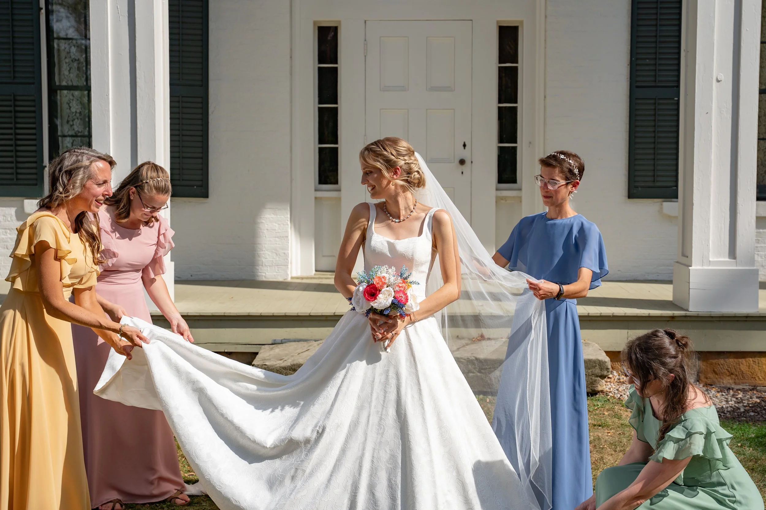 A bride in a white wedding dress holding a bouquet, standing outside a white house with green shutters, surrounded by five women in colorful dresses adjusting her veil and dress.