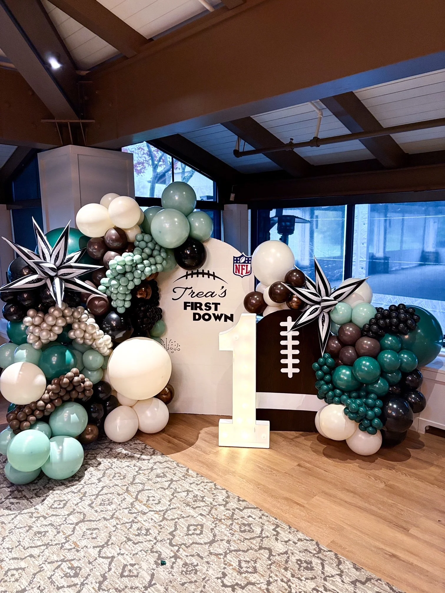 Decorative balloon arrangement with football theme, featuring a large number 1, a football graphic, and signs reading "Trea's First Down" for a first birthday celebration.
