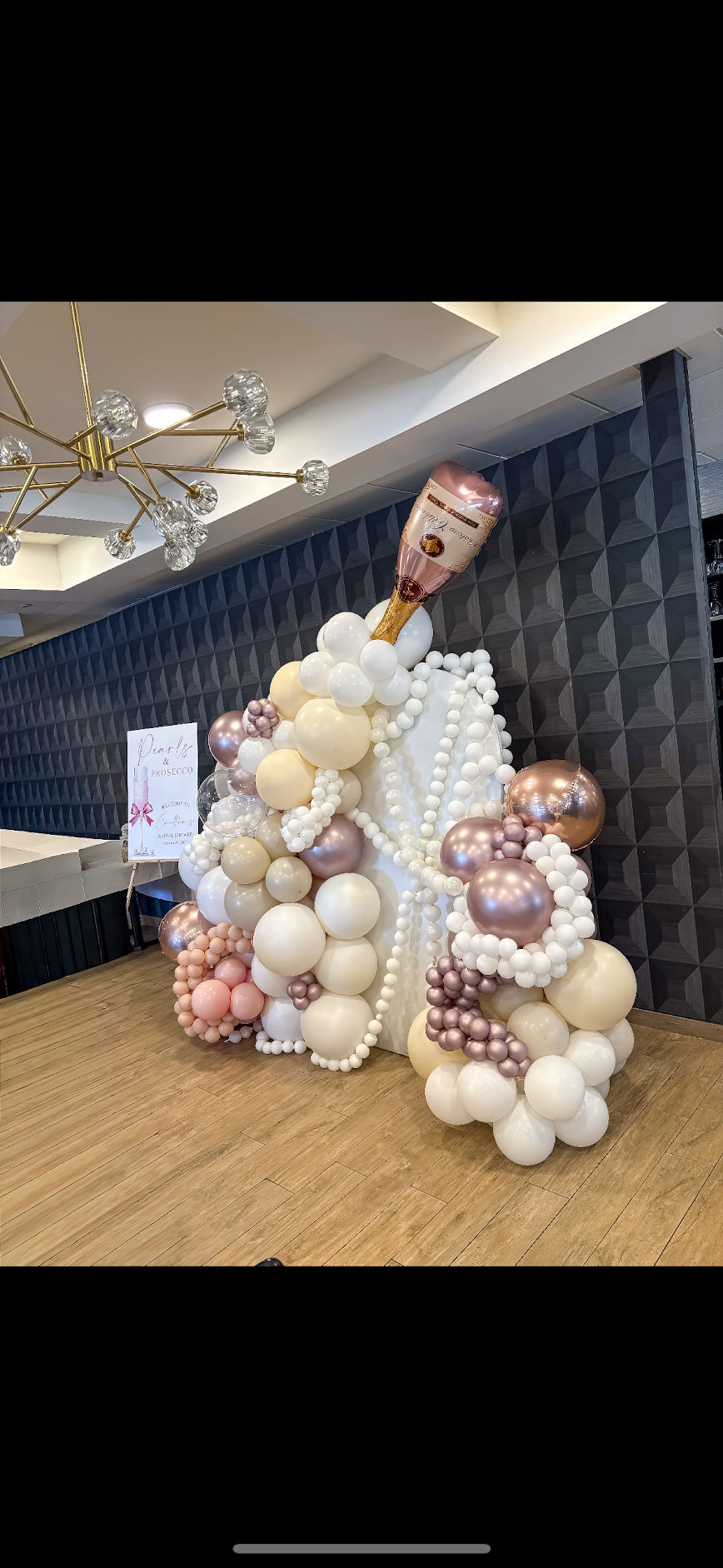 Balloon display with a large champagne bottle balloon on top, arranged with variously sized white, pink, peach, and metallic balloons, set against a black textured wall with a gold chandelier overhead.