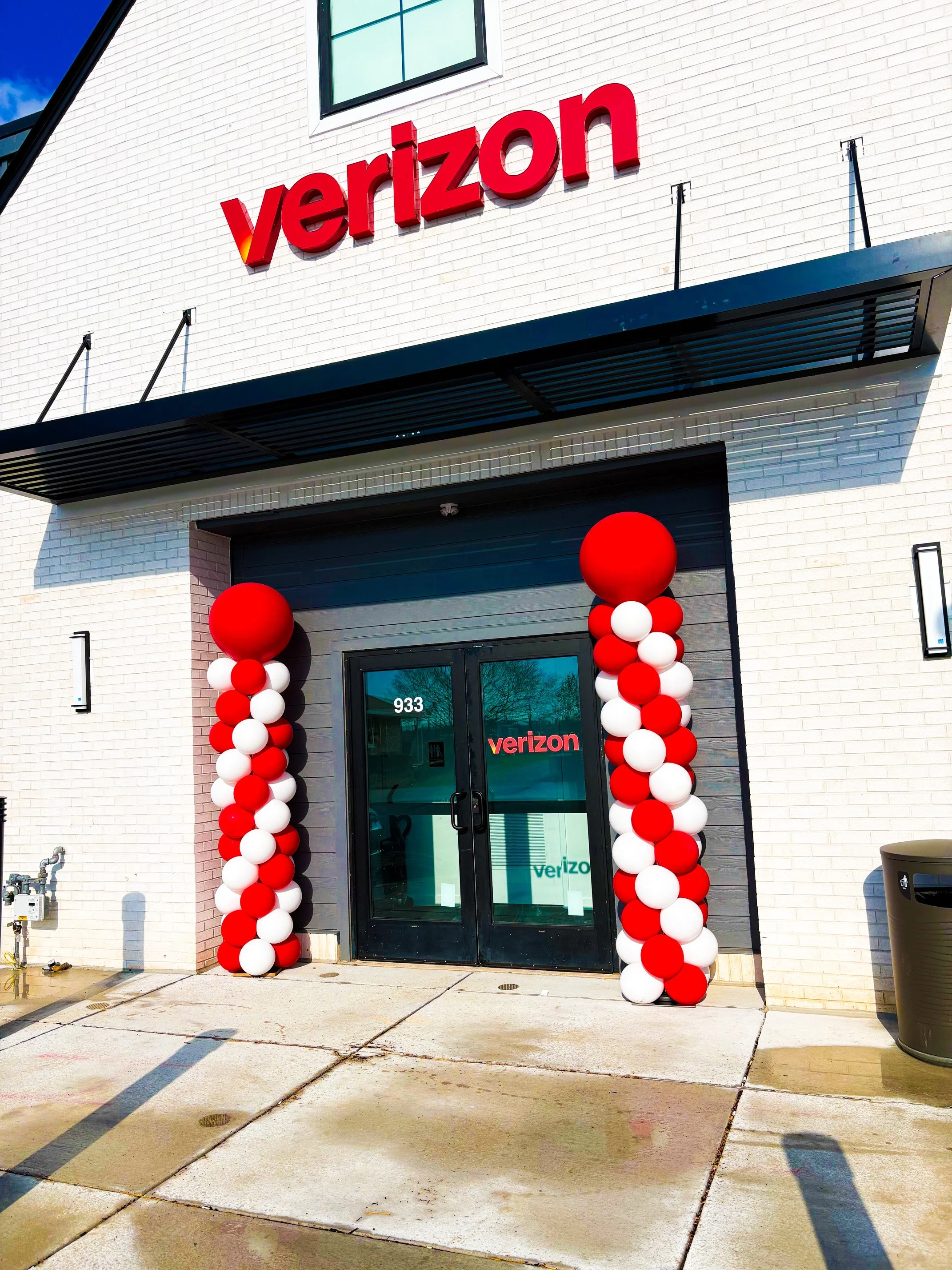 Verizon store entrance decorated with red and white balloon columns, with the Verizon logo above the door and on the glass doors.