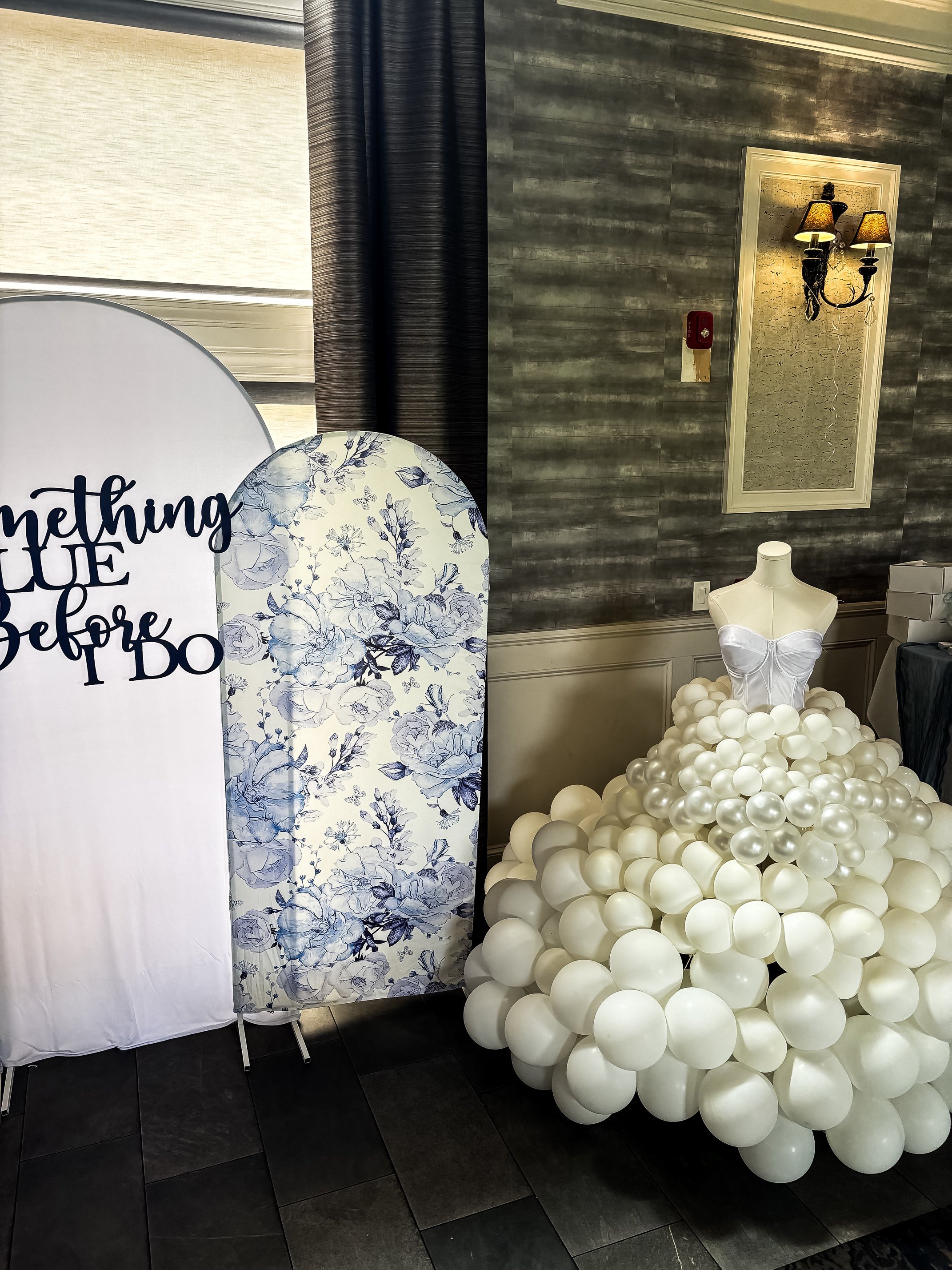 A decorative display with a mannequin wearing a white corset top set among numerous white balloons forming a skirt. To the left, there are two fabric-covered panels; one with floral and the other with text that reads 'something blue before'. The back