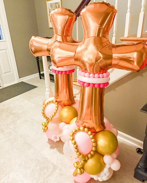 Decorative balloon arrangement with large rose gold number balloons, gold and pink balloons, and pearl-like rings, placed indoors near a staircase and wall.