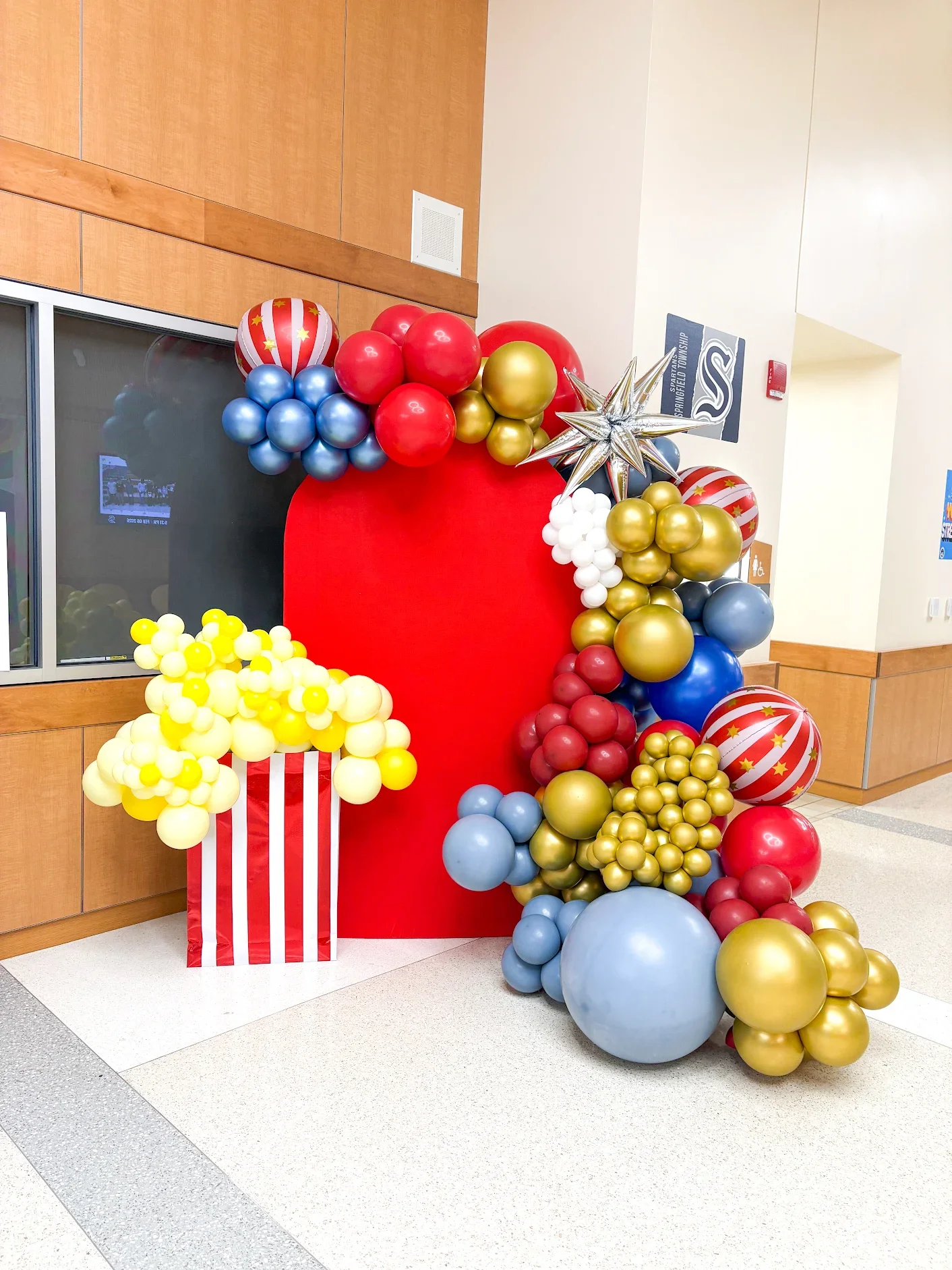Decorative balloon arrangement with red, blue, gold, and white balloons, some with stars and stripes, arranged around a large red backdrop and a striped paper container, in an indoor lobby area.