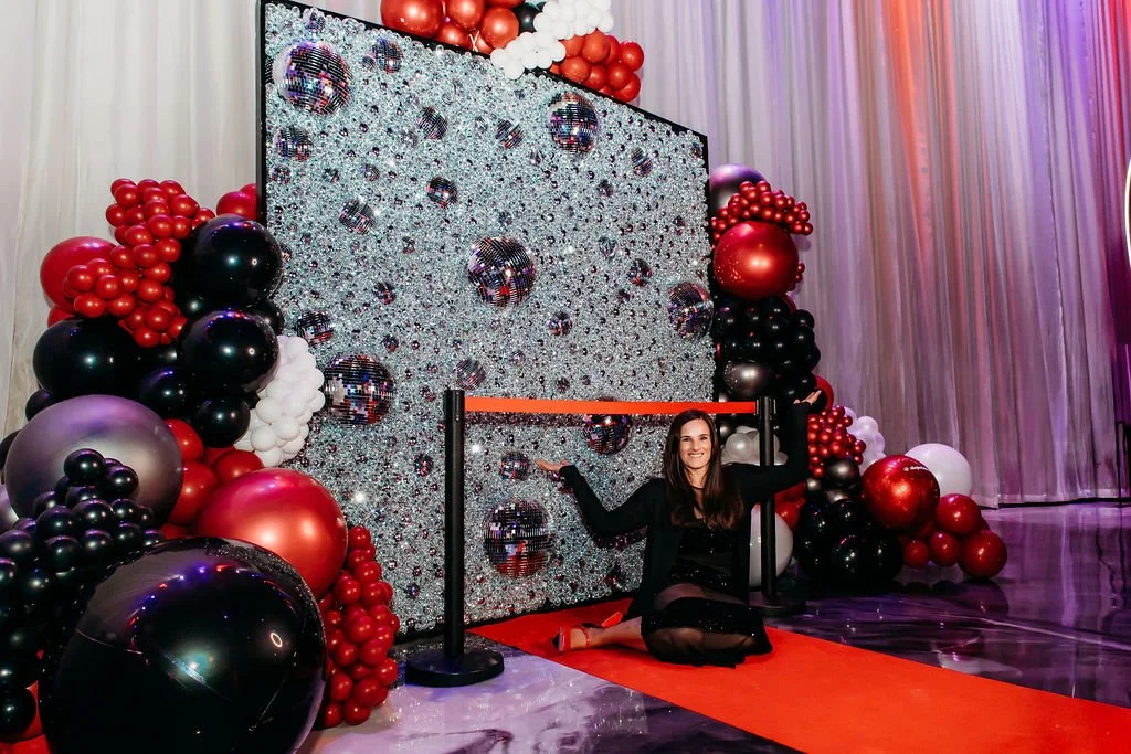 A woman sitting on a red carpet in front of a decorated backdrop with balloons and disco balls at a celebration event.
