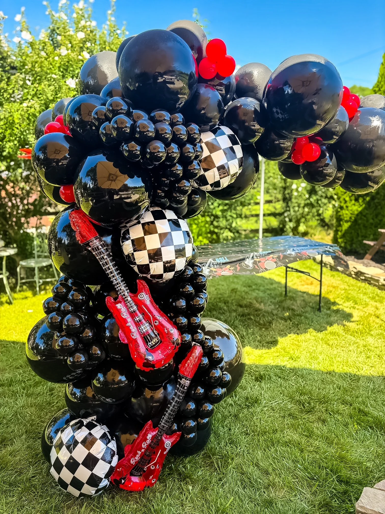 A large balloon sculpture of a guitar made with black, red, and white checkered balloons, situated outdoors on green grass with trees and a blue sky in the background.