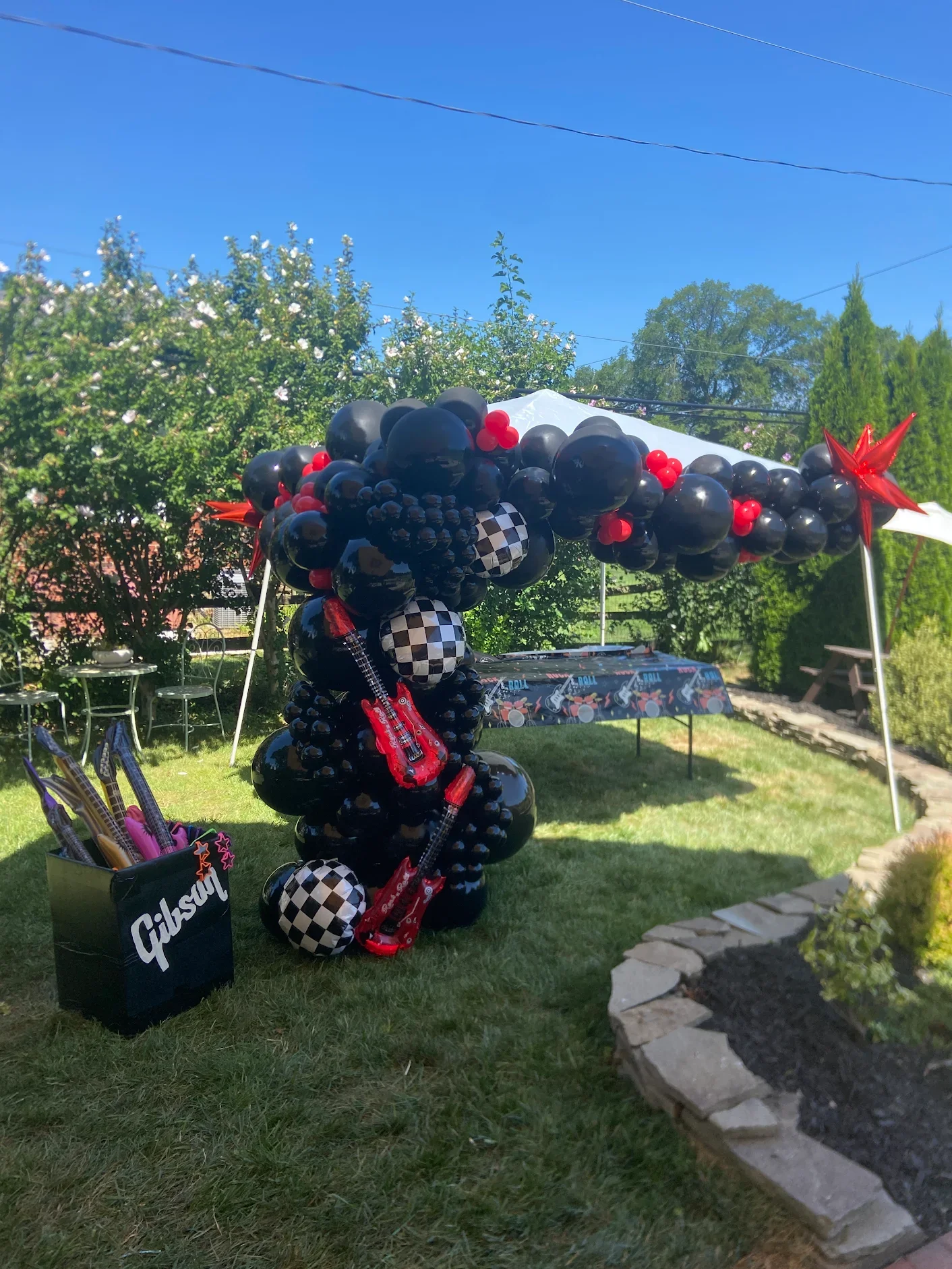 Outdoor birthday party setup with black and red balloons, and guitar decorations under a canopy, with a table and chairs in the background on a sunny day.