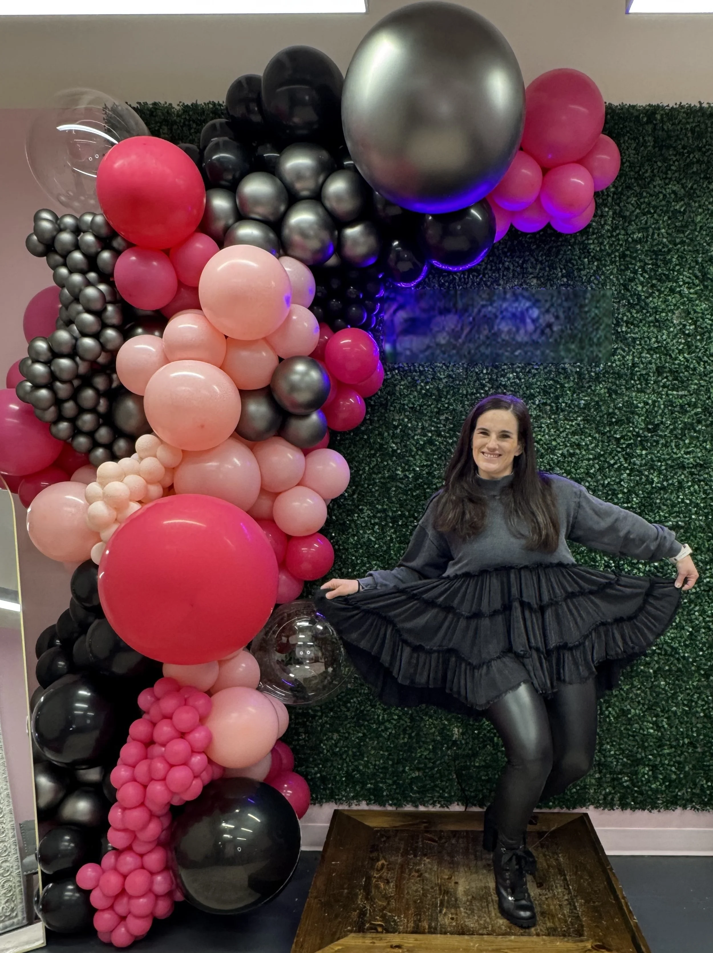 A woman stands in front of a wall of colorful balloons arranged in an arch and cluster, smiling and holding the edge of her black layered skirt, with a green foliage background.