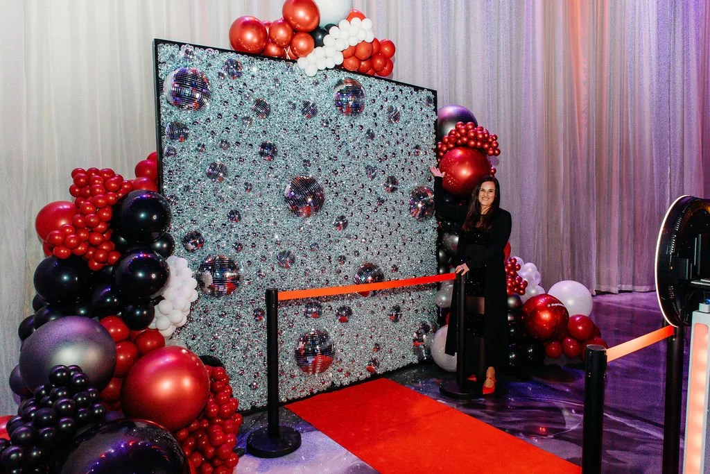 A woman standing next to a starry disco-themed photo backdrop decorated with red, black, white, and purple balloons, with a red carpet and a velvet rope barrier in front.
