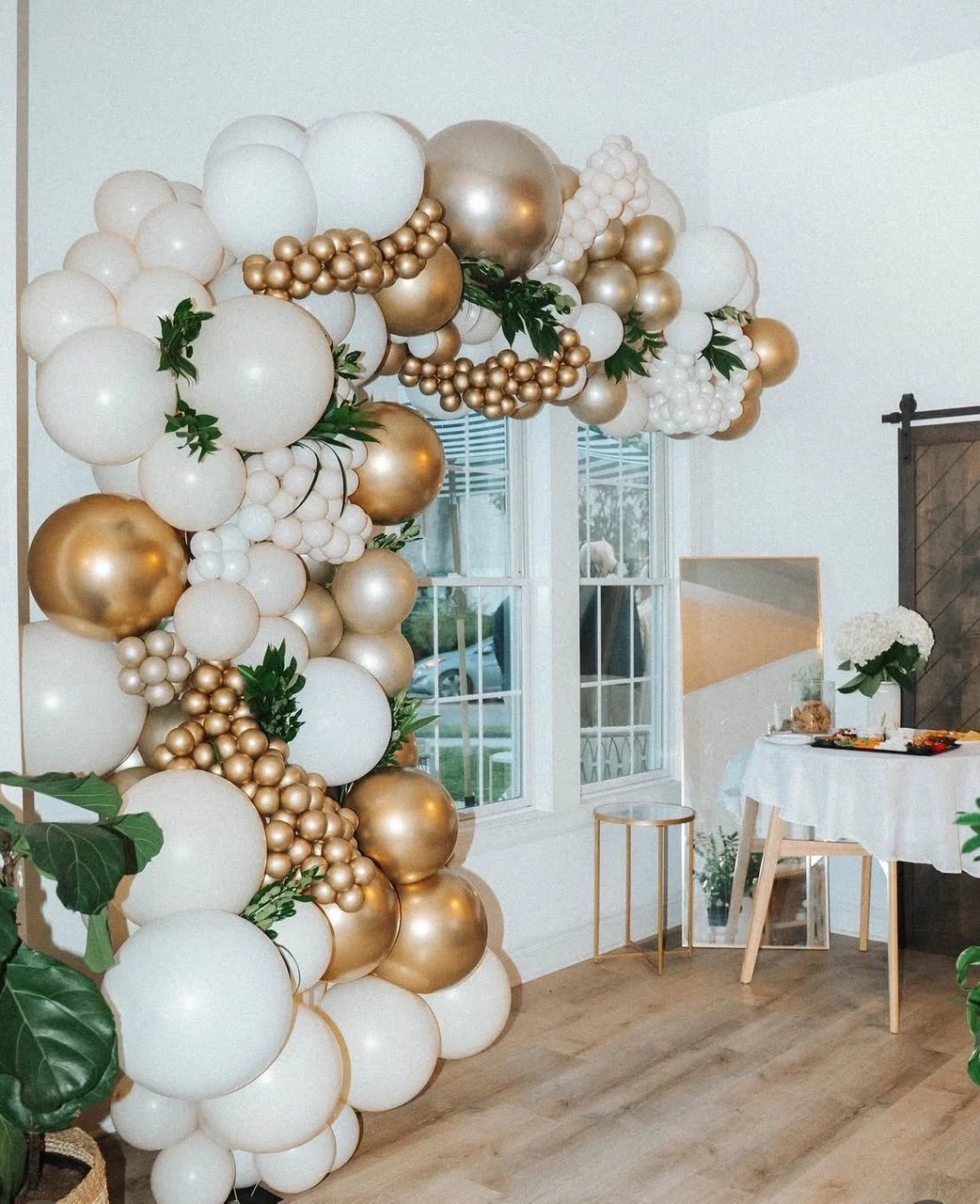 Balloon display with white, gold, and champagne balloons, decorated with green leaves, in a room with a window, a mirror, and a table with a white tablecloth and floral accents.