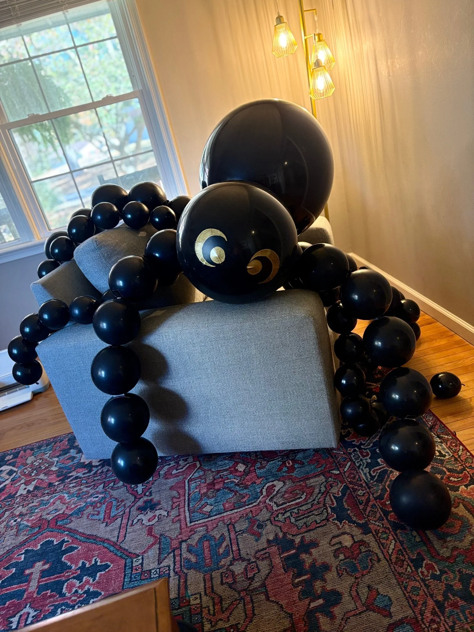 Decorative arrangement of large black balloons with gold accents on a gray couch in a living room with a colorful area rug and a window showing trees outside.