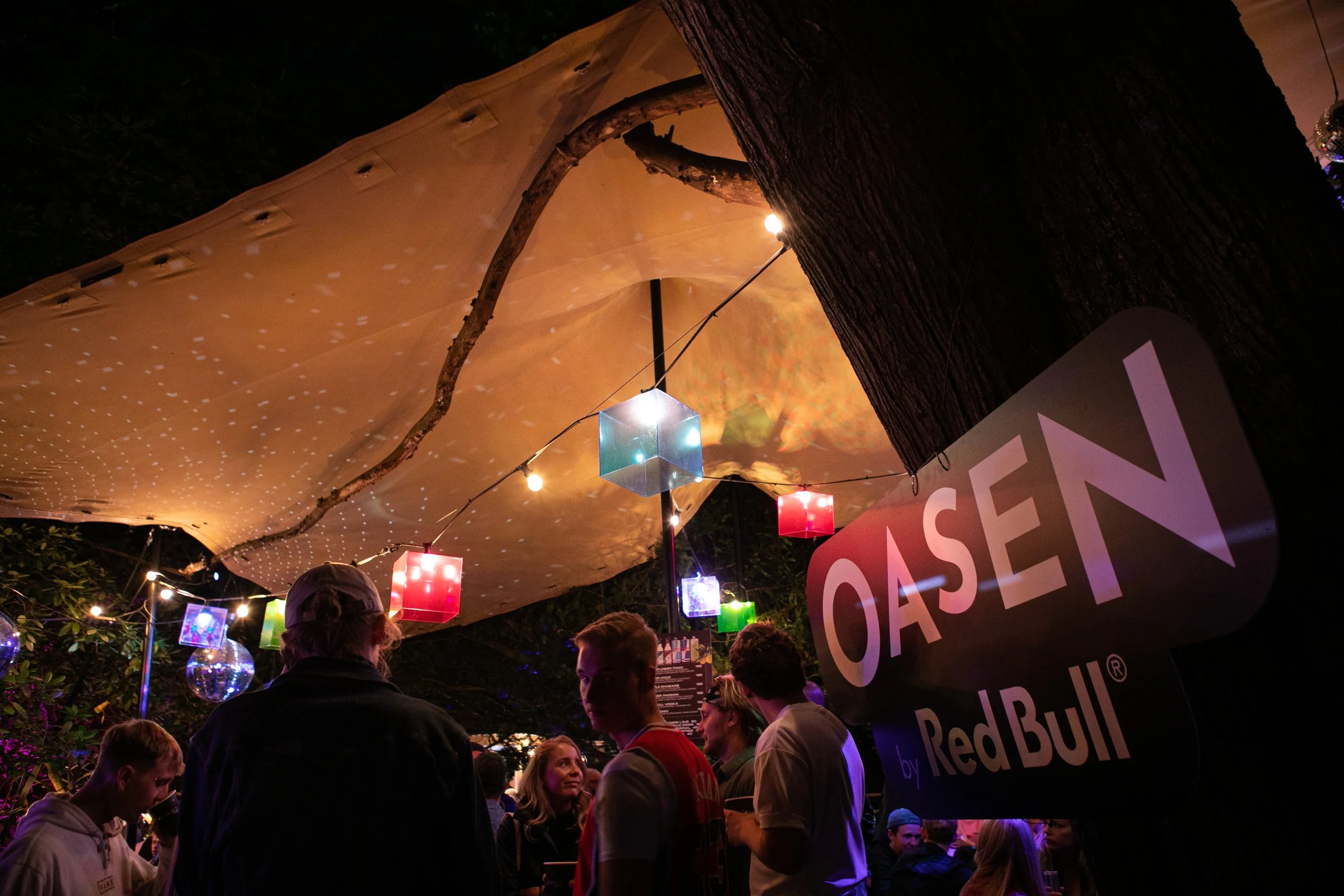 Människor samlas under en utomhusbiograf med färgglada lampor och ett vävt tygdraperi, nära ett träd, vid en Red Bull- och Oasen-anläggning.