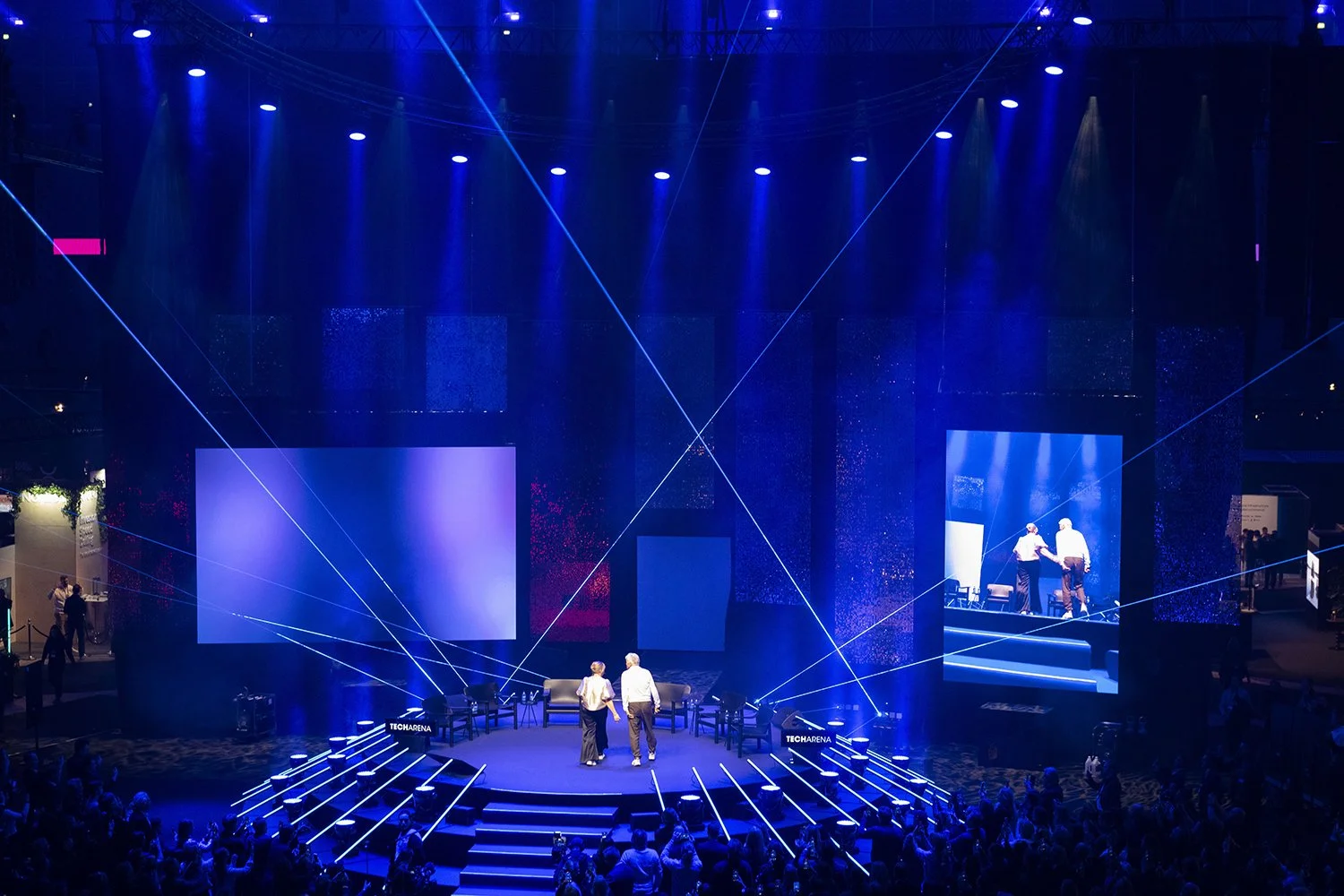 Tech Arena Scen med två personer på en scen med blå belysning och flera skärmar, omgivande publik.