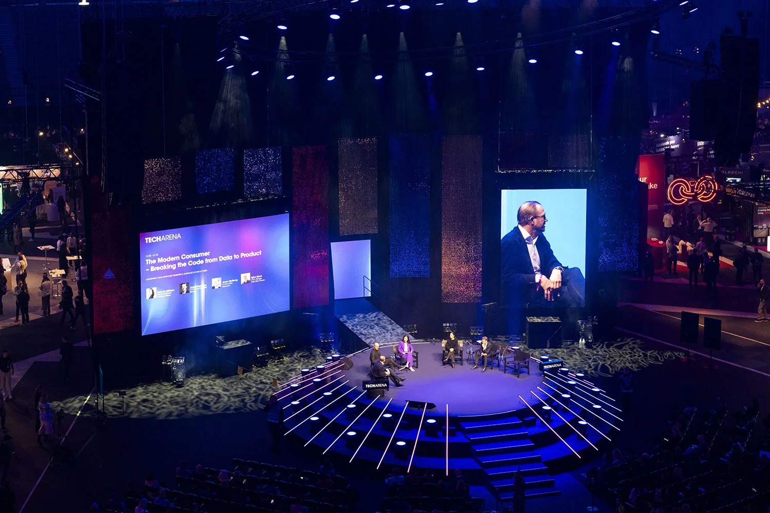En scen med ett panelavsnitt på TechArena-konferensen, med fem personer som pratar och ett stort skärminlägg med texten 'The Modern Consumer - Breaking the Code from Data to Product', omgivet av färgglada ljus och publiken.
