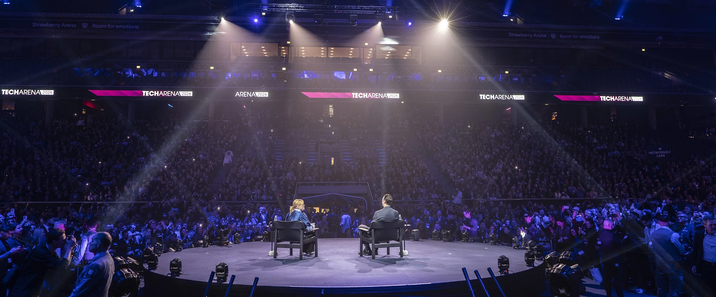 En stor publik i en arena med två personer som pratar på en scen, upplyst av spotlight, med digitala skärmar som visar 'TECHARENA 2026' och 'Room for emotions'.