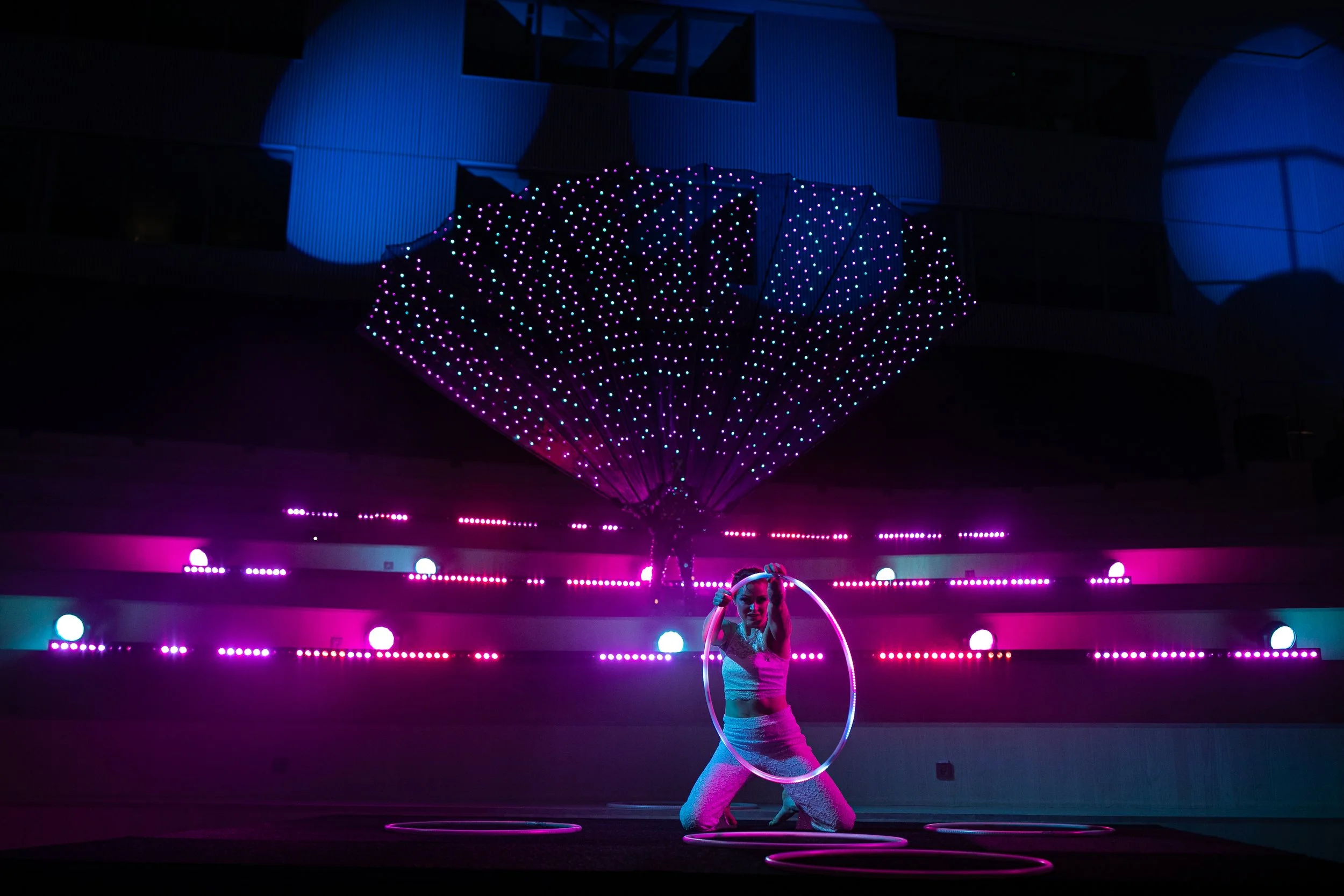 En människa som jonglerar med ljusbollar i en mörk miljö med färgglada LED-lampor och en stor, upplyst strukturerad kupol i bakgrunden.