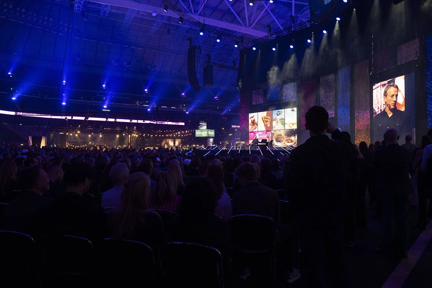 En stor publik sitter och tittar på en scenduk under ett evenemang i en inomhushall med blå och lila belysning och flera skärmar.