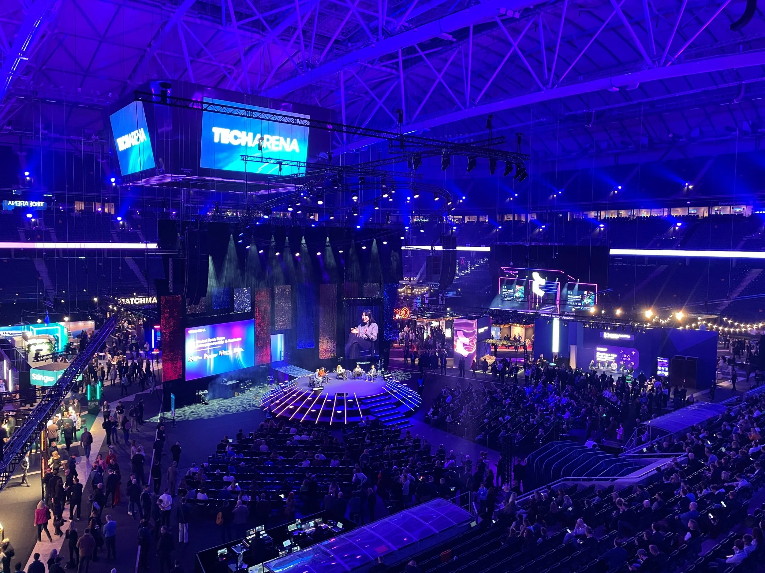 En stor inomhusarena med en scen och många publikmedlemmar, upplyst av blå och lila belysning.