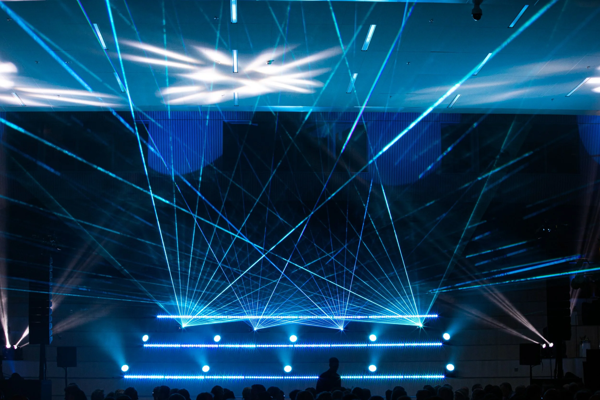 Laserljus- och ljusevenemang i en mörk sal, med blå och vita ljusstrålar och skuggor av personer i publiken.