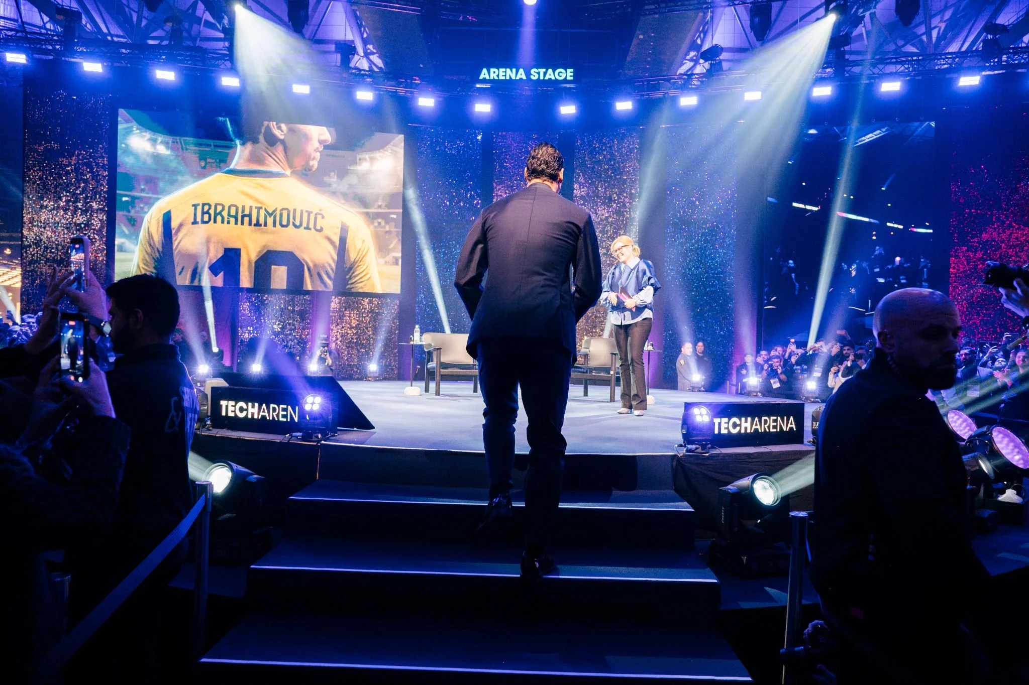 En scen med många människor, ett stort skärmbanskt med en person i en gul sporttröja med namnet Ibrahimovic, och en man som går uppför en trappa mot scenen med texter 'Tech Arena' och 'Arena Stage' ovanför. Officiella evenemanget verkar vara teknolog