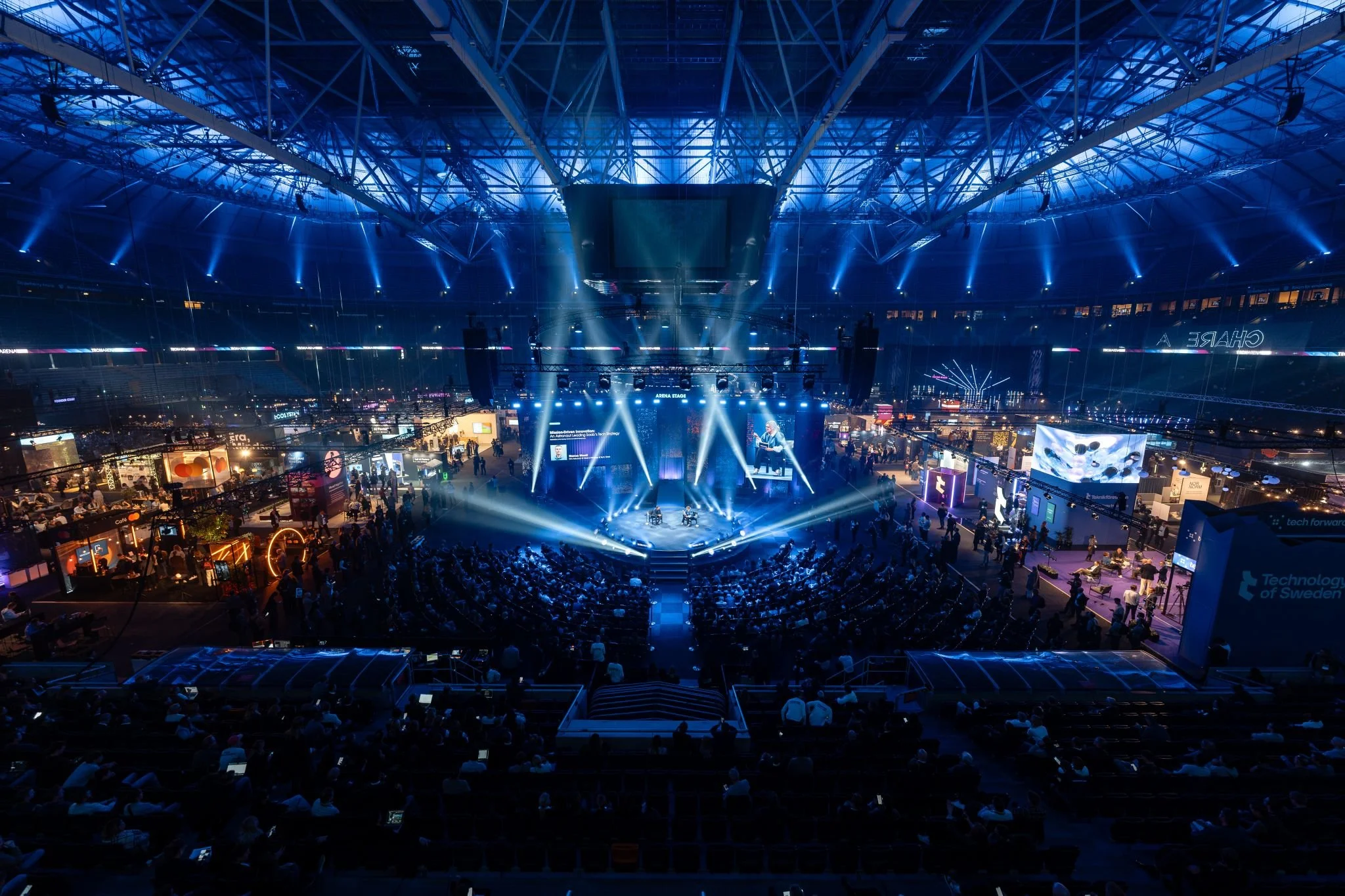 Stort inomhusevent med ett scengalleri med flera personer på scenen, cirkulerande ljus och publik i sittplatser framför scenen, omgiven av olika utställningsstånd med människor, i en stor modern hall med glasdome och blå belysning.