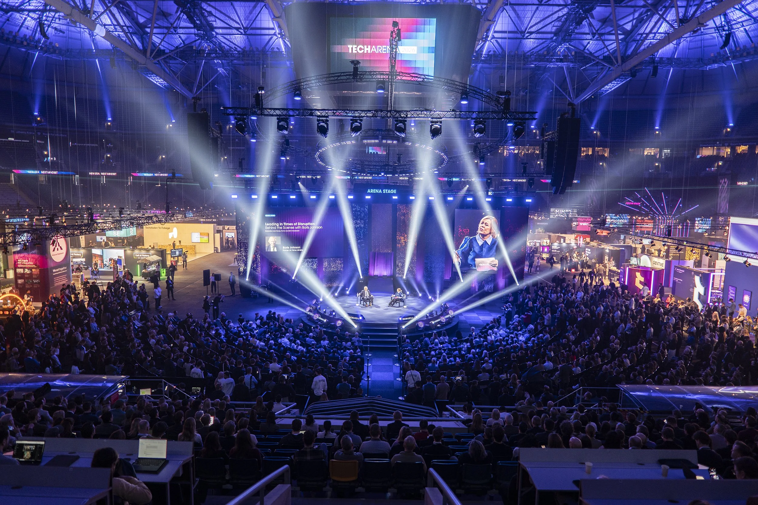 Tech Arena Stort publik eventsarena med scen och två personer som talar, omgiven av ljus- och videoprojektioner och utställningar.