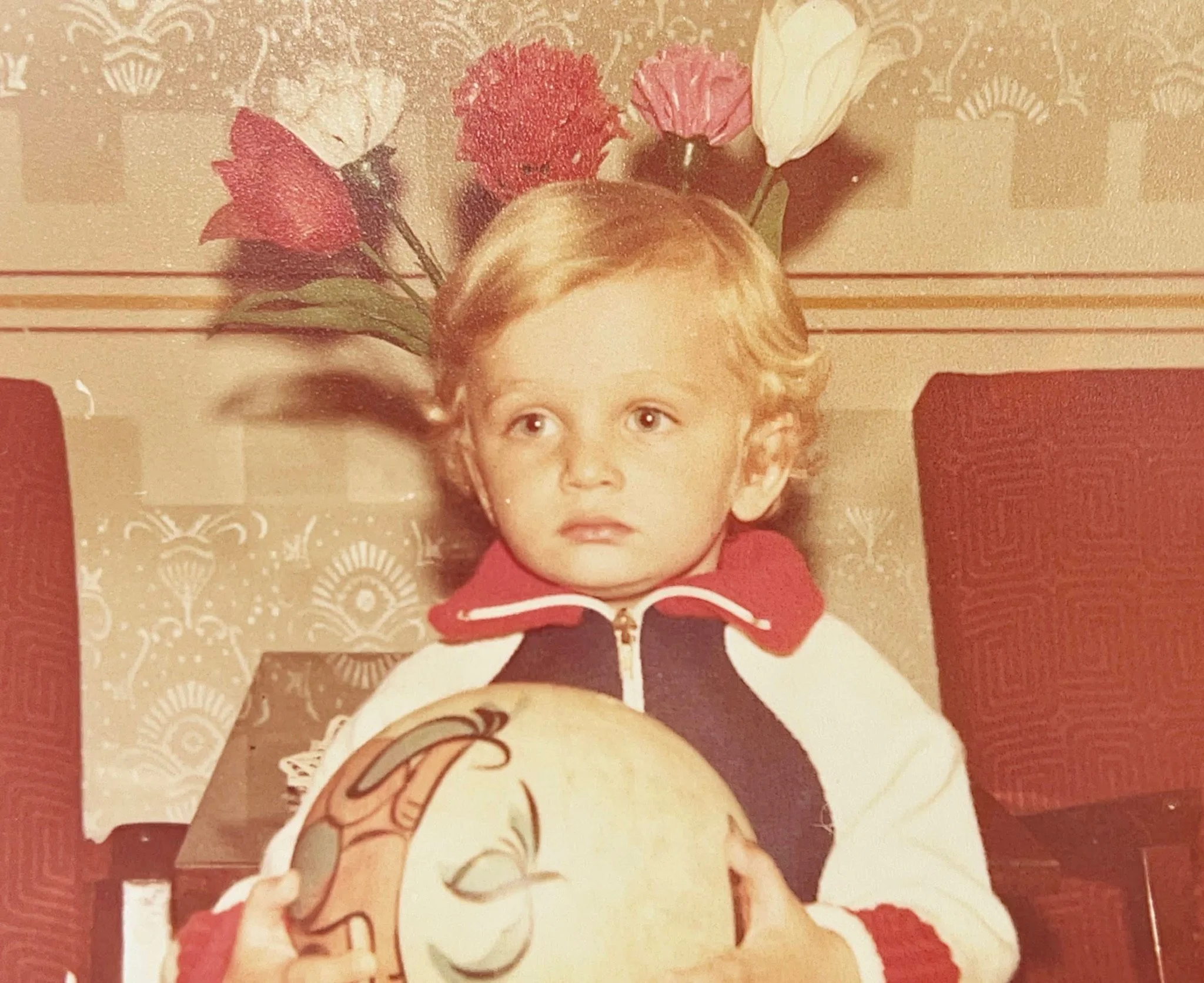 A young boy with curly blonde hair sitting at a dining table, holding a decorated balloon, with pink and white flowers in the background.