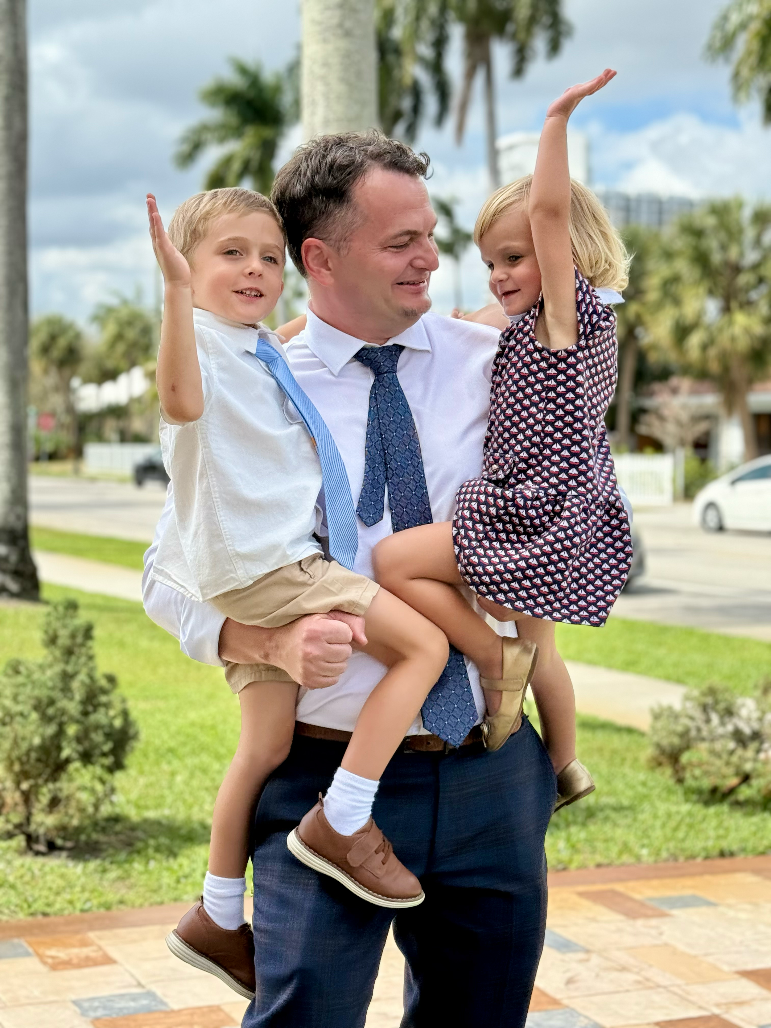 A man in a white dress shirt and blue tie holding two children, a boy in a white shirt and khaki shorts and a girl in a patterned dress, both with their arms raised, outdoors with palm trees and cars in the background.