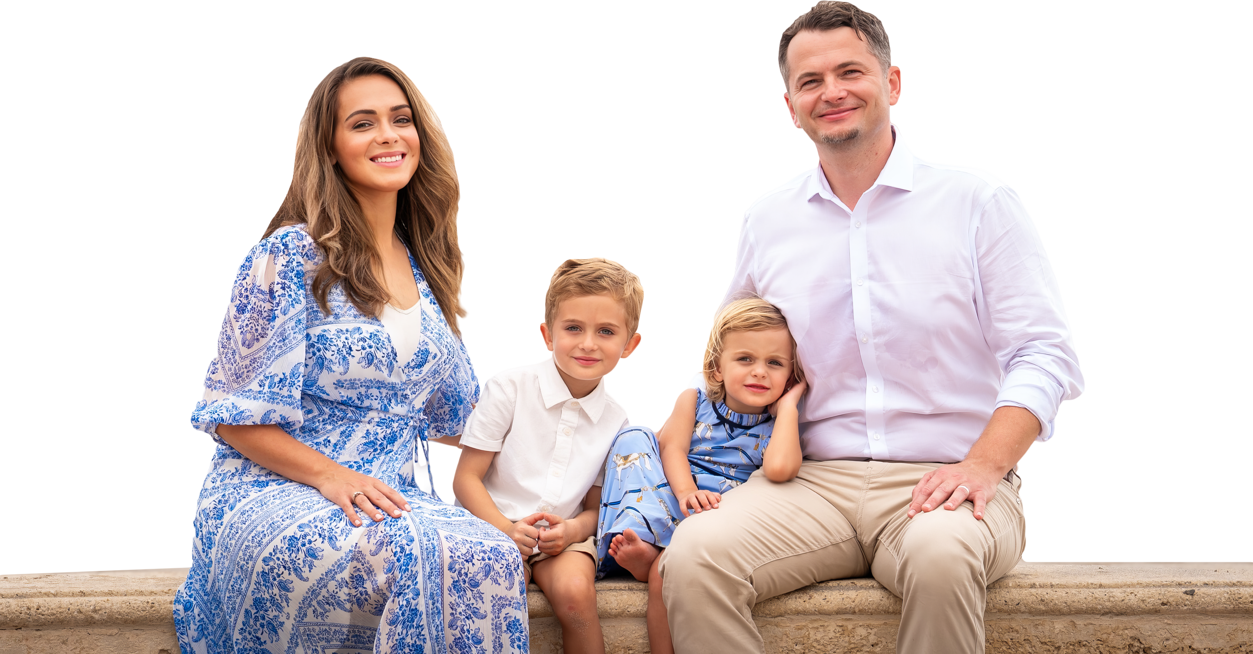 A family of five sitting on a stone ledge outdoors, smiling. The family includes a woman wearing a blue and white dress, a man in a white shirt and beige pants, and three children—a boy and a girl, all dressed in light-colored clothing.