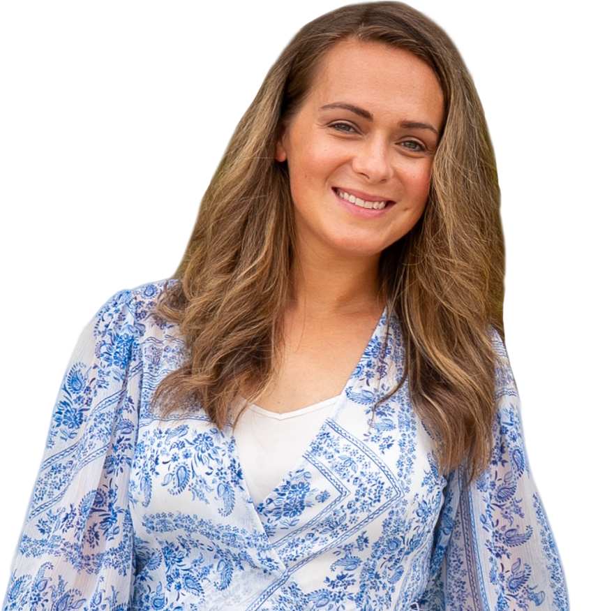 A smiling woman with long, wavy brown hair wearing a white and blue floral patterned blouse, standing outdoors.