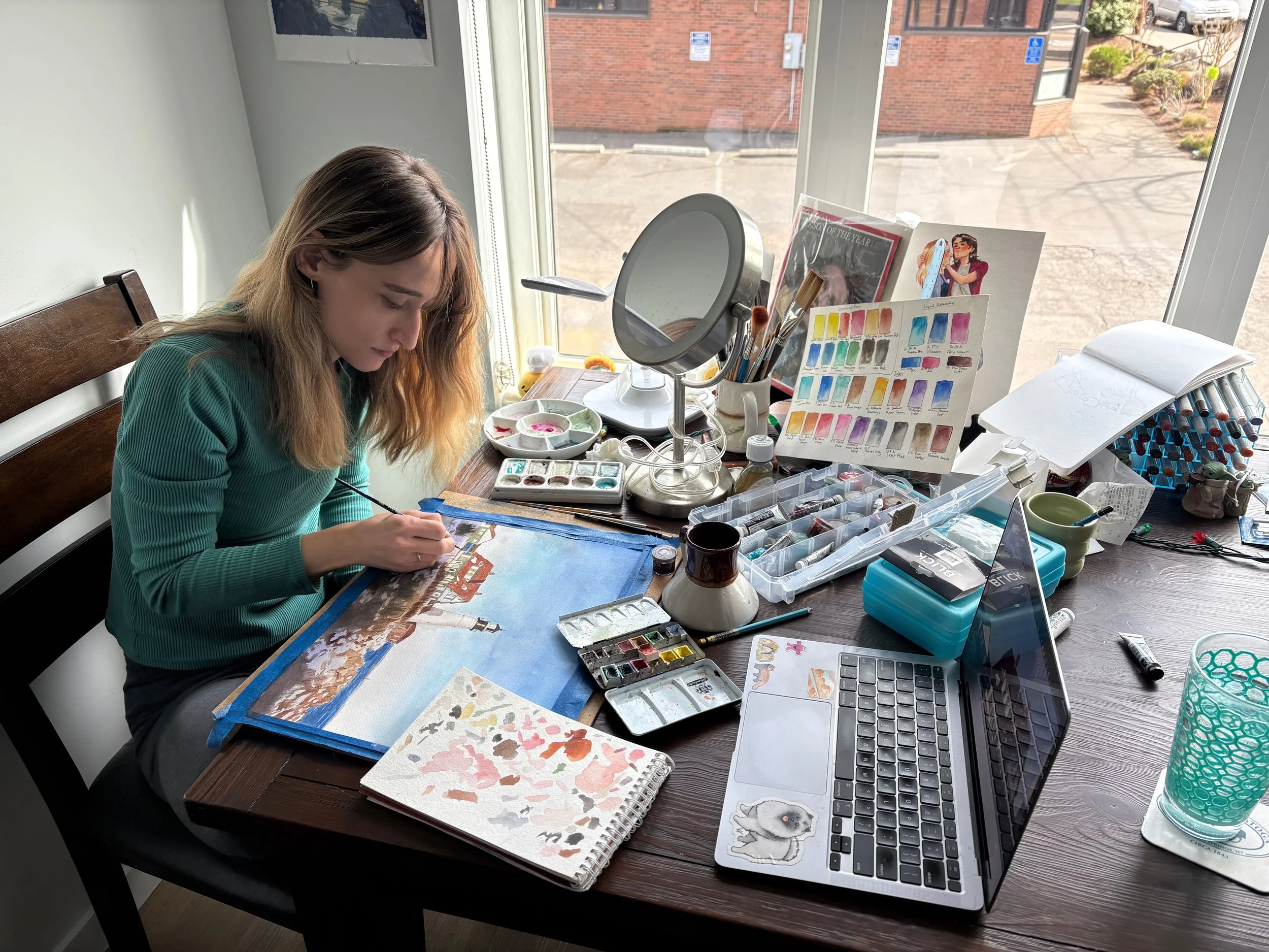 Watercolor artist and painter painting a watercolour work of Portland, Maine. It is the Cape Elizabeth lighthouse. Ocean view with blue sky, snow, at sunset.