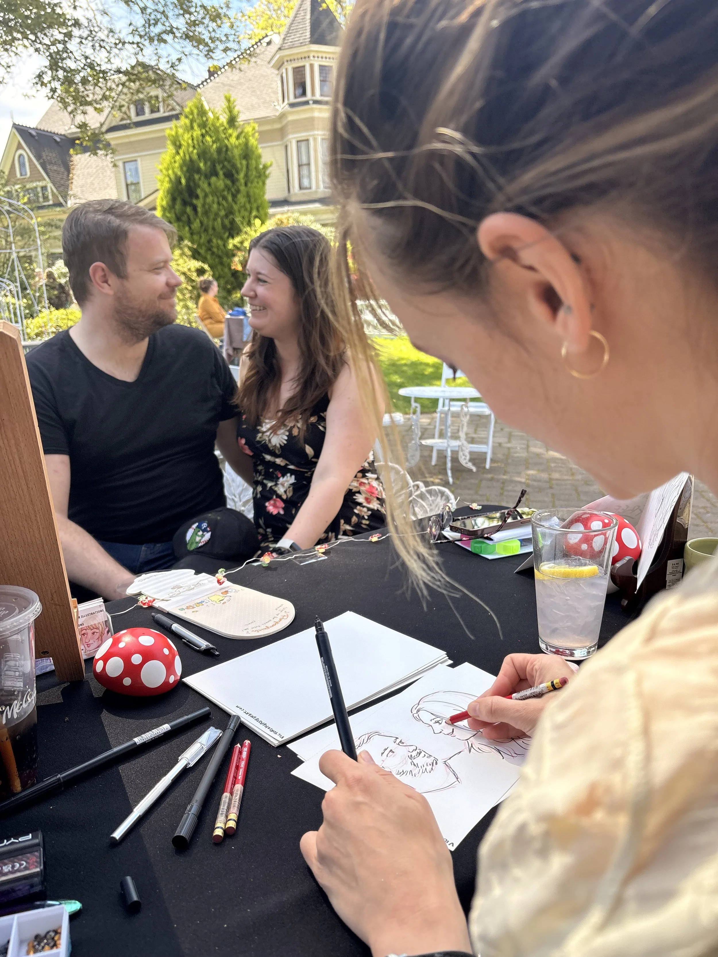 A live portrait artists draws an engaged couple sitting for a live portrait drawing at a wedding expo. They are all outside and the Victorian Belle mansion house is in the background.