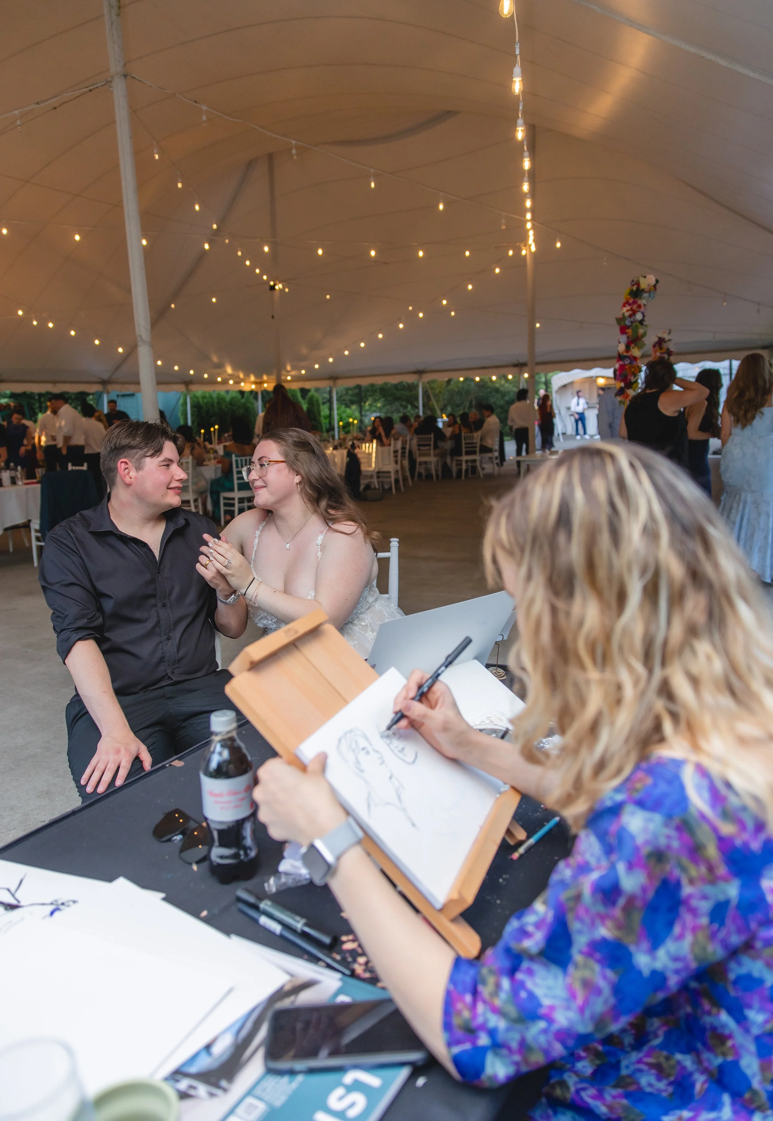 A couple sits at a table during a wedding celebration under a large tent with string lights. The bride and groom are looking at each other and holding hands, while a portrait artist sketches them.