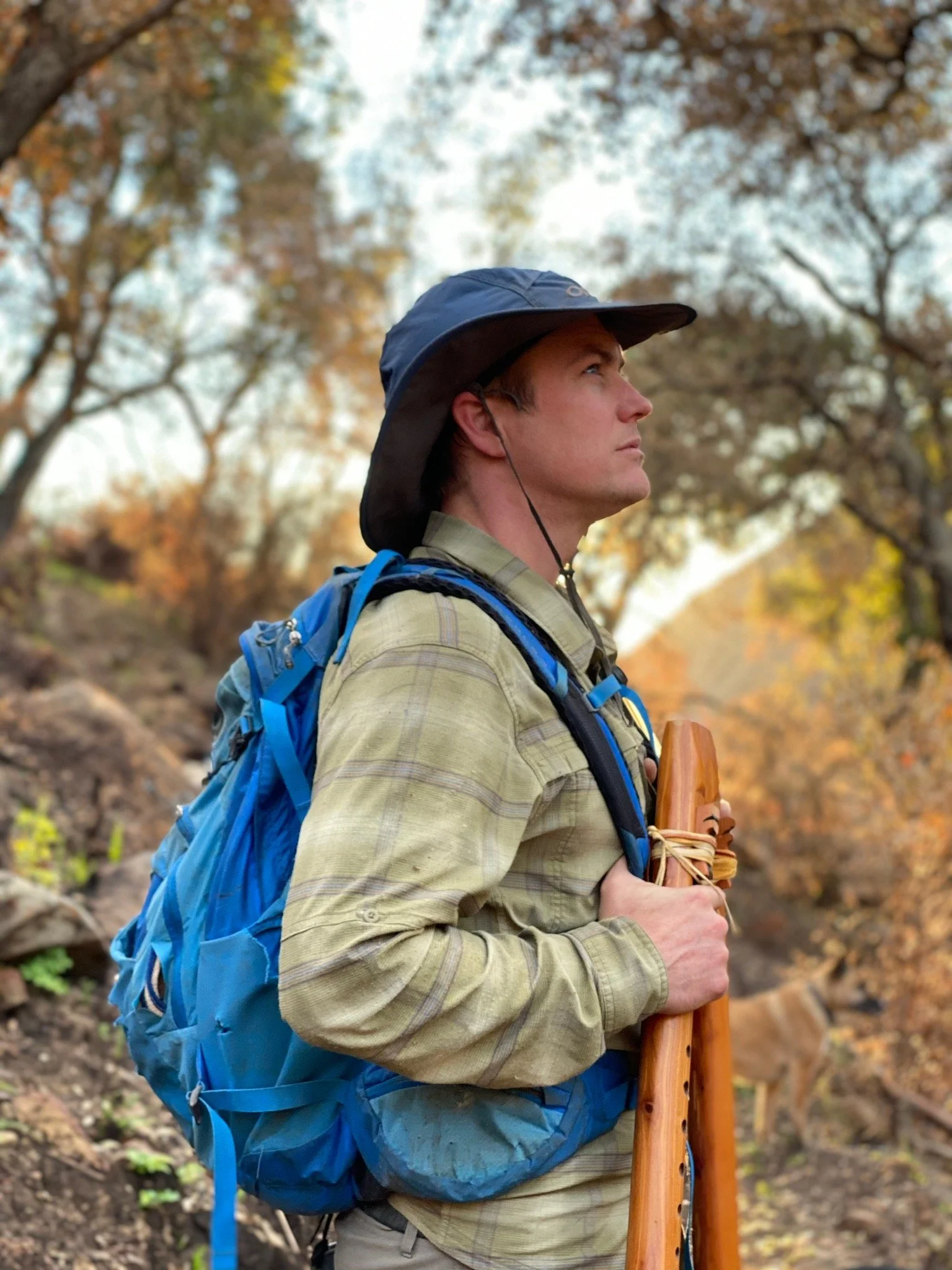 A man wearing a wide-brimmed hat and a plaid shirt stands outdoors on a trail surrounded by autumn trees, holding a wooden walking stick and wearing a blue backpack.