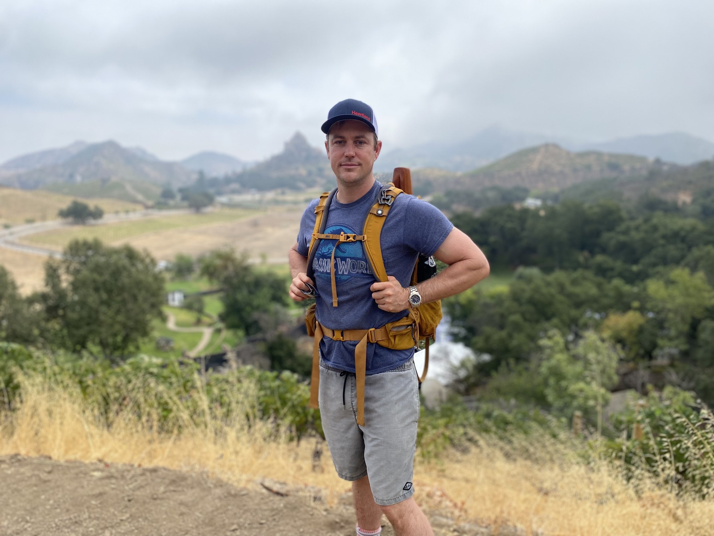 A man stands outdoors on a hiking trail with a backpack, wearing a baseball cap and casual clothing, surrounded by a landscape of rolling hills and cloudy sky.