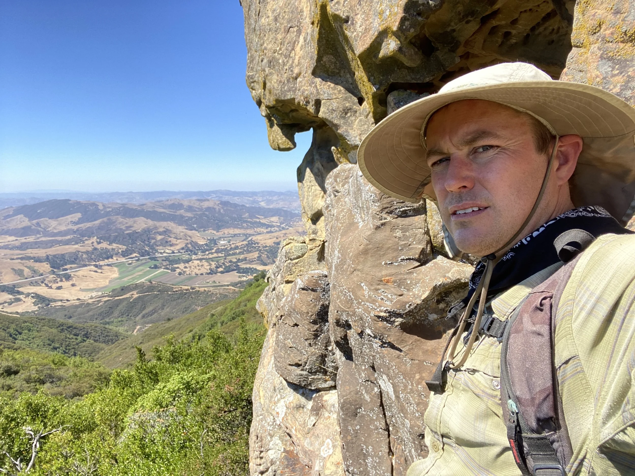 A man wearing a large beige hat and outdoor gear stands against a rocky mountain ledge overlooking a valley with rolling hills, fields, and distant mountains under a clear blue sky.