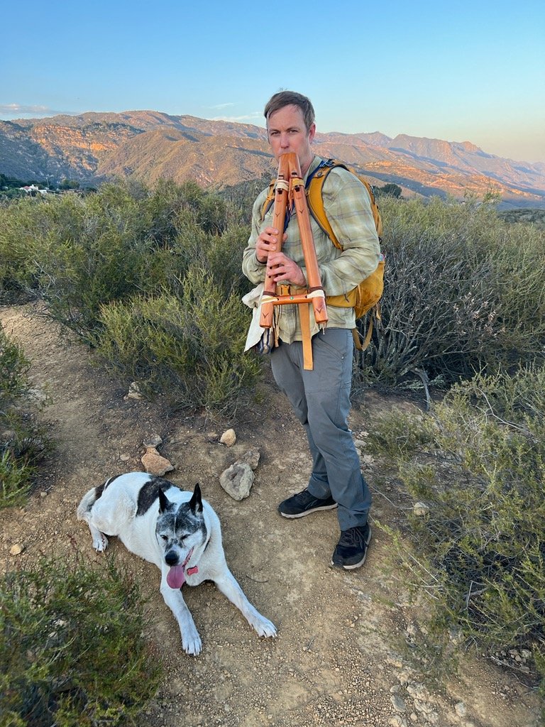 A man playing a wooden pan flute with a backpack standing on a dirt trail in a mountainous area, accompanied by a white and black dog, during sunset or early evening.
