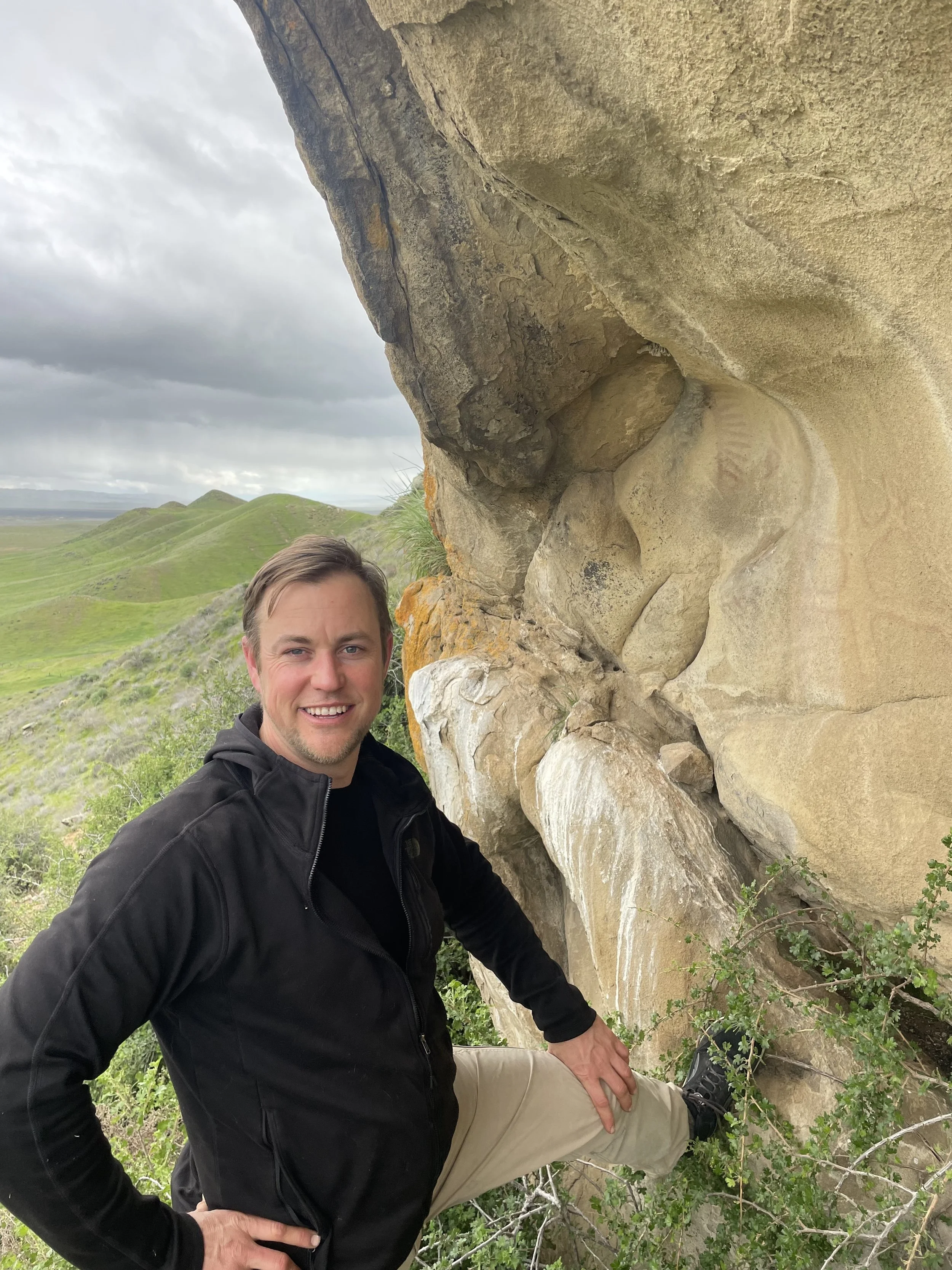 A man with short brown hair, wearing a black jacket and beige pants, posing outdoors next to a large rock formation with a cloudy sky and green rolling hills in the background.