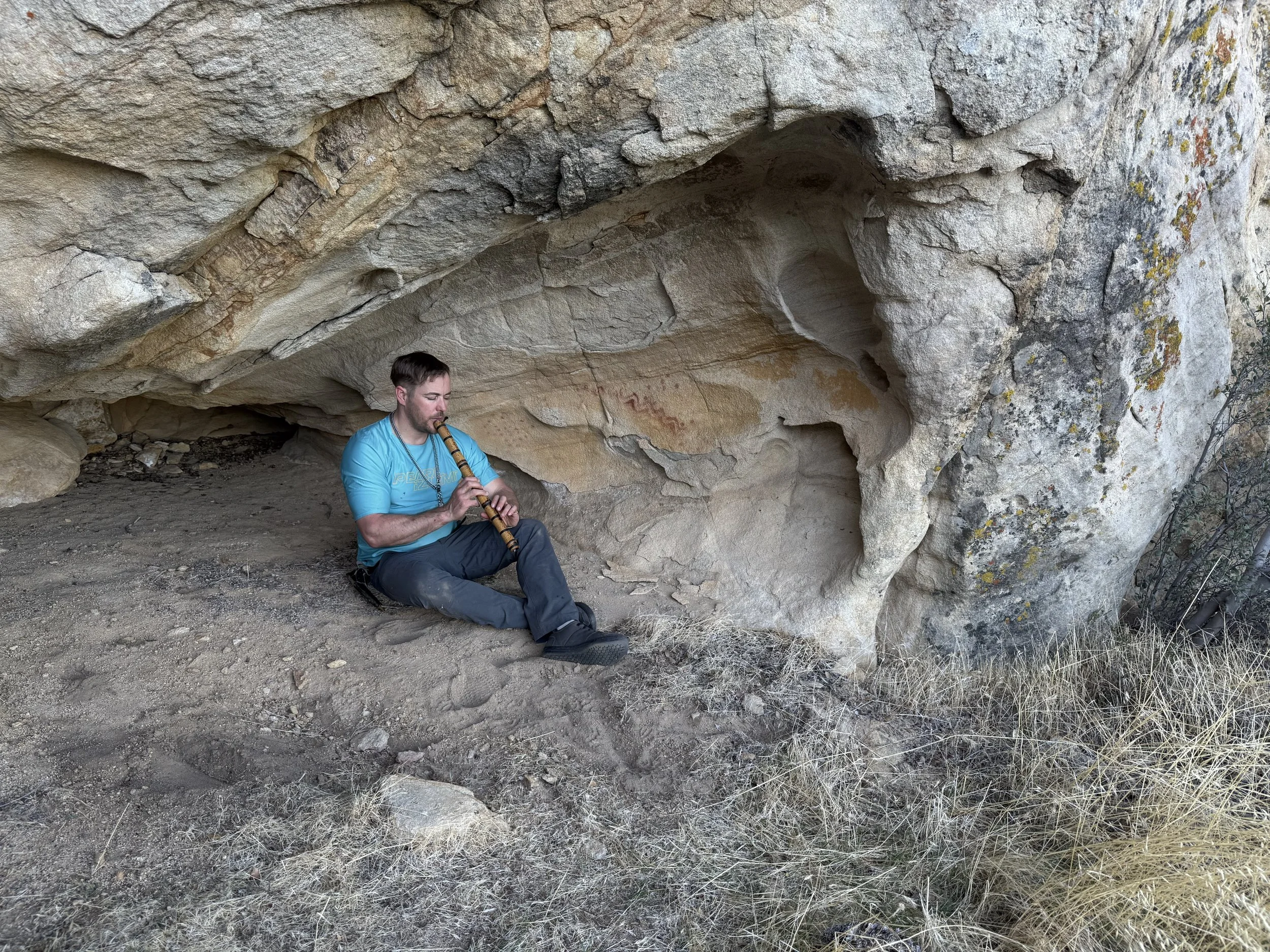 A man sitting on the ground under a rocky overhang, playing a wooden flute, in a natural outdoor setting with dry grass and rocks.