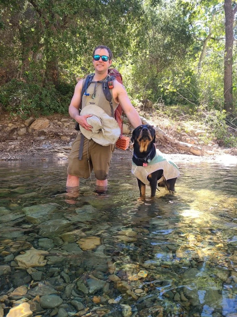 A man and a dog standing in a shallow creek surrounded by trees. The man is wearing sunglasses, a tan tank top, shorts, and a backpack, holding a wide-brimmed hat. The dog is a black and tan breed wearing a light-colored vest.