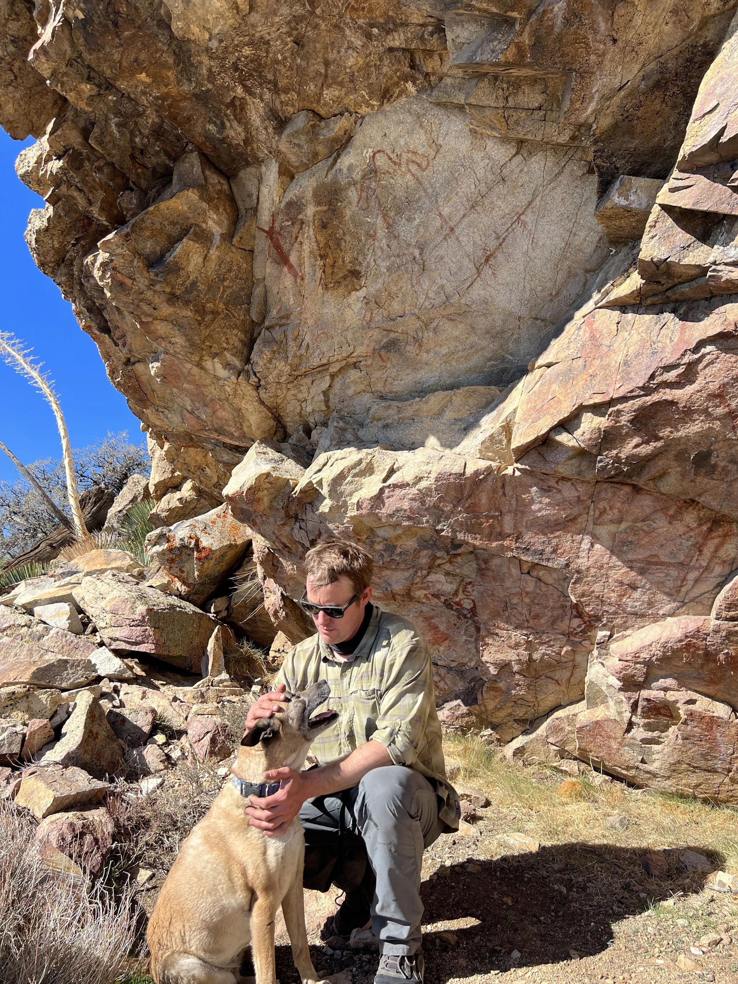 A man in outdoor gear is kneeling and petting a large dog with a tan coat and black facial markings, while sitting on rocky ground in front of a large rock formation with petroglyphs carved into it. The scene is outdoors with a clear blue sky and spa