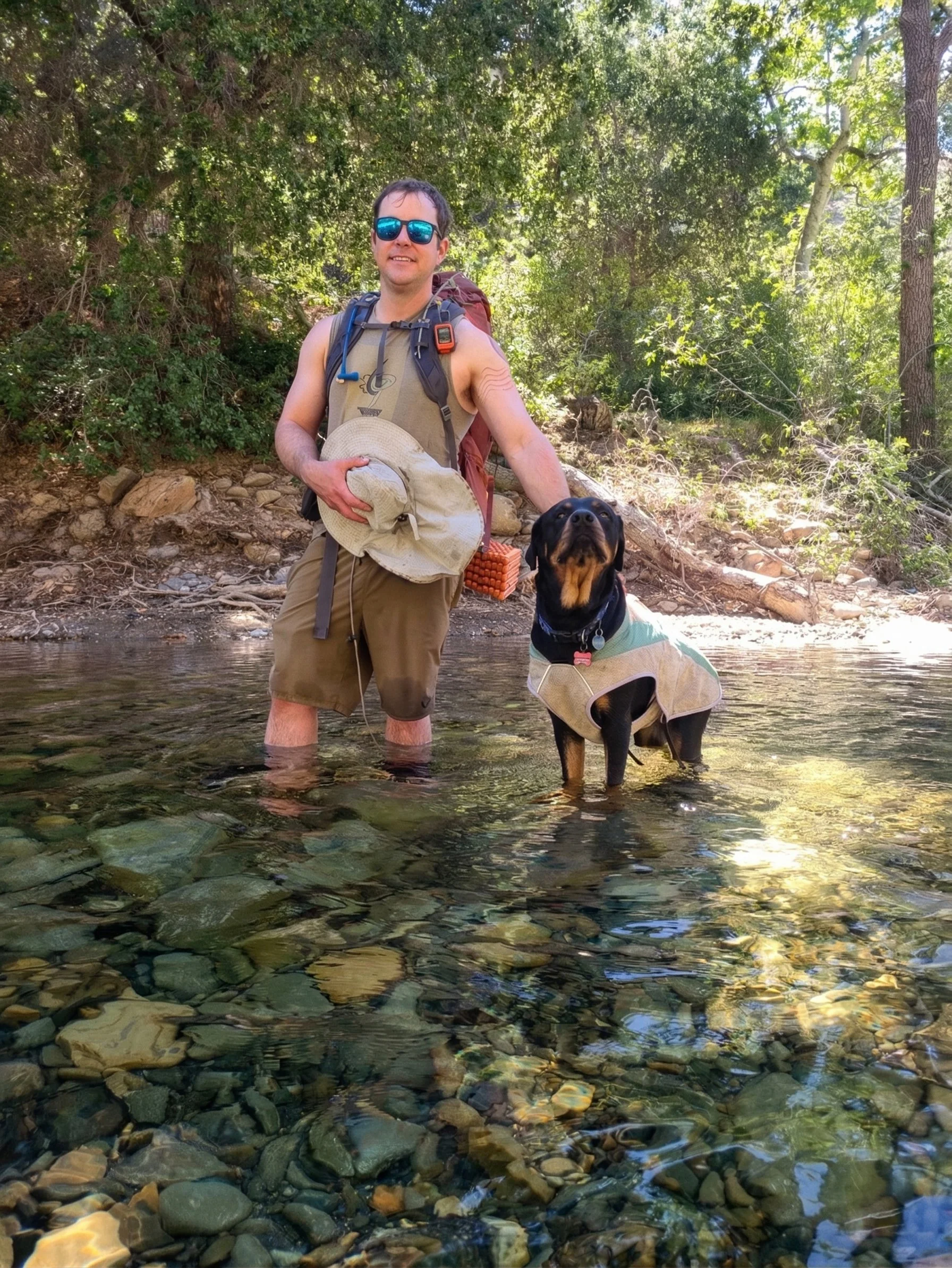 Eric Stokell wearing sunglasses, a sleeveless shirt, and hiking gear stands in a stream with a dog wearing a vest, surrounded by lush green trees and rocks.