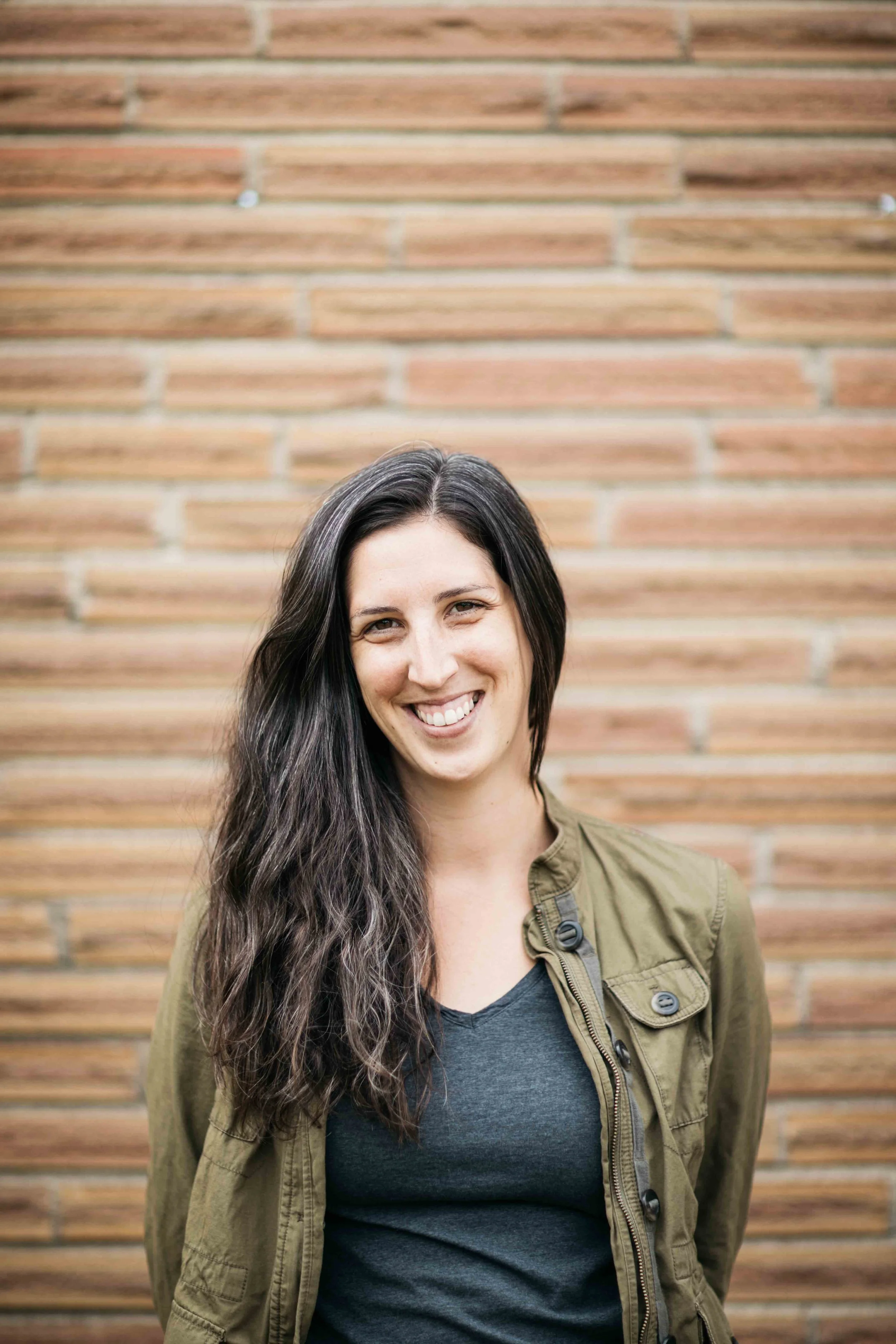 A woman with long, wavy dark hair smiling and standing in front of a brick wall, wearing an olive-green jacket and a dark t-shirt.