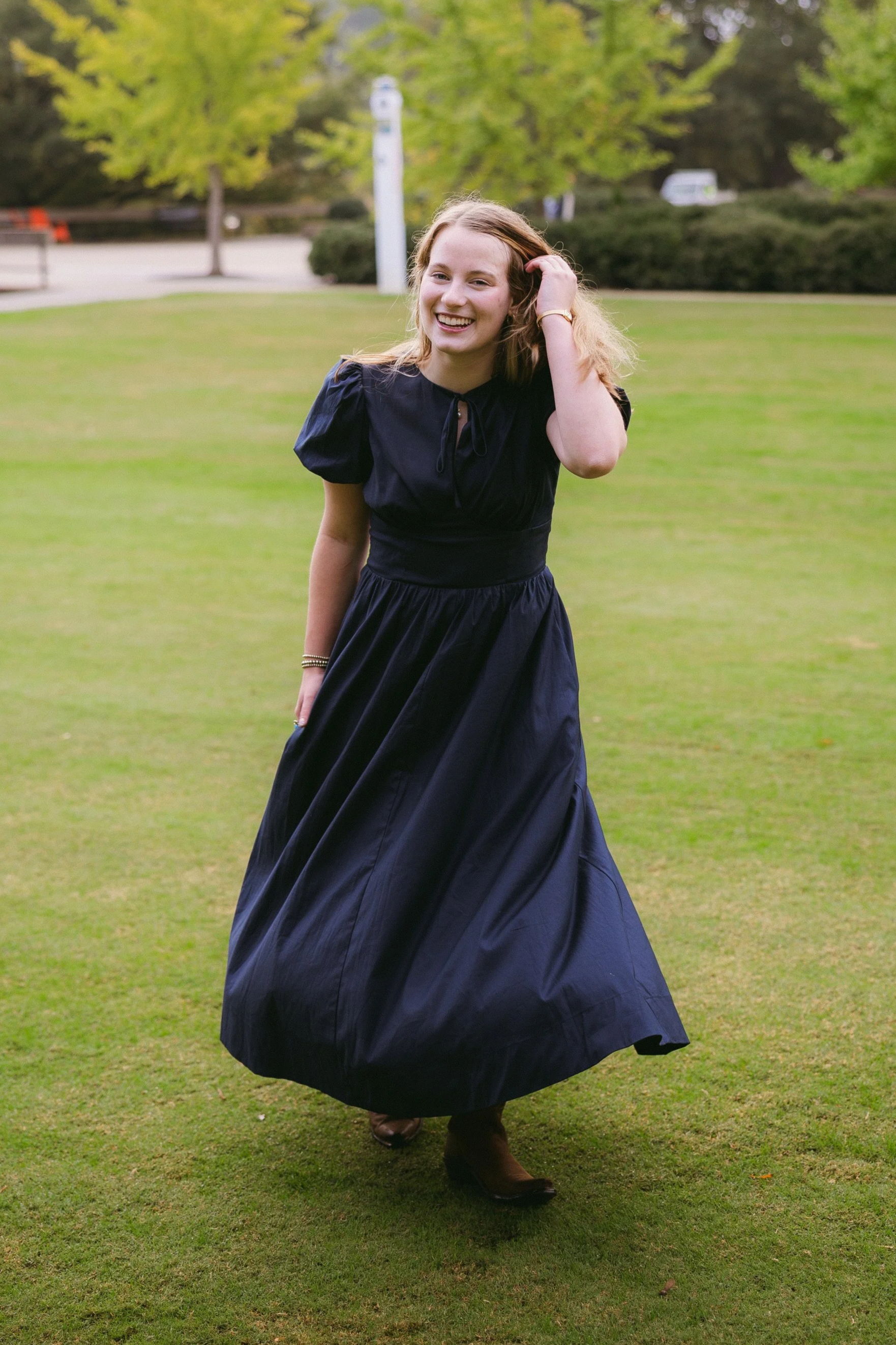 A young woman wearing a long, dark blue dress with puffed sleeves, smiling and running on a grassy field with trees and a parking lot in the background.